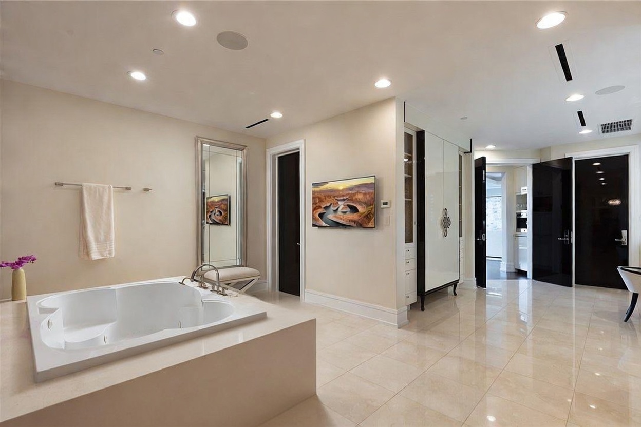 This spacious primary bathroom features a large, built-in soaking tub as the focal point, complemented by a tall, framed mirror and a wall-mounted television. The room is finished with light-toned tile flooring and neutral wall colors, creating a bright and airy atmosphere. A doorway leads to an adjacent area, while a sleek, dark-toned door adds a modern contrast to the clean, minimalist aesthetic.