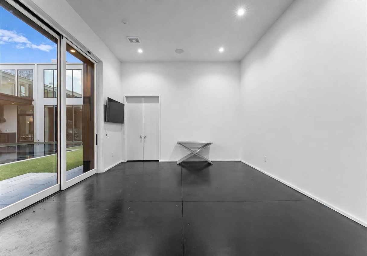 This minimalist interior space features dark, polished concrete flooring and stark white walls, creating a clean and modern aesthetic. A large sliding glass door provides a seamless transition to an outdoor pool area, while a small metallic console table sits against the back wall. The room is illuminated by recessed lighting and offers a bright, open atmosphere.