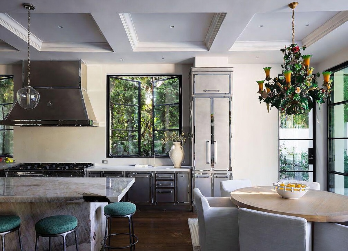 This sophisticated kitchen features a large stone-topped island with green upholstered stools and a professional-grade range with a prominent stainless steel hood. The space is accented by a decorative floral chandelier over a round dining table and dark metal cabinetry that contrasts with the light-toned walls. Large windows provide a view of lush greenery, creating a bright and elegant atmosphere.