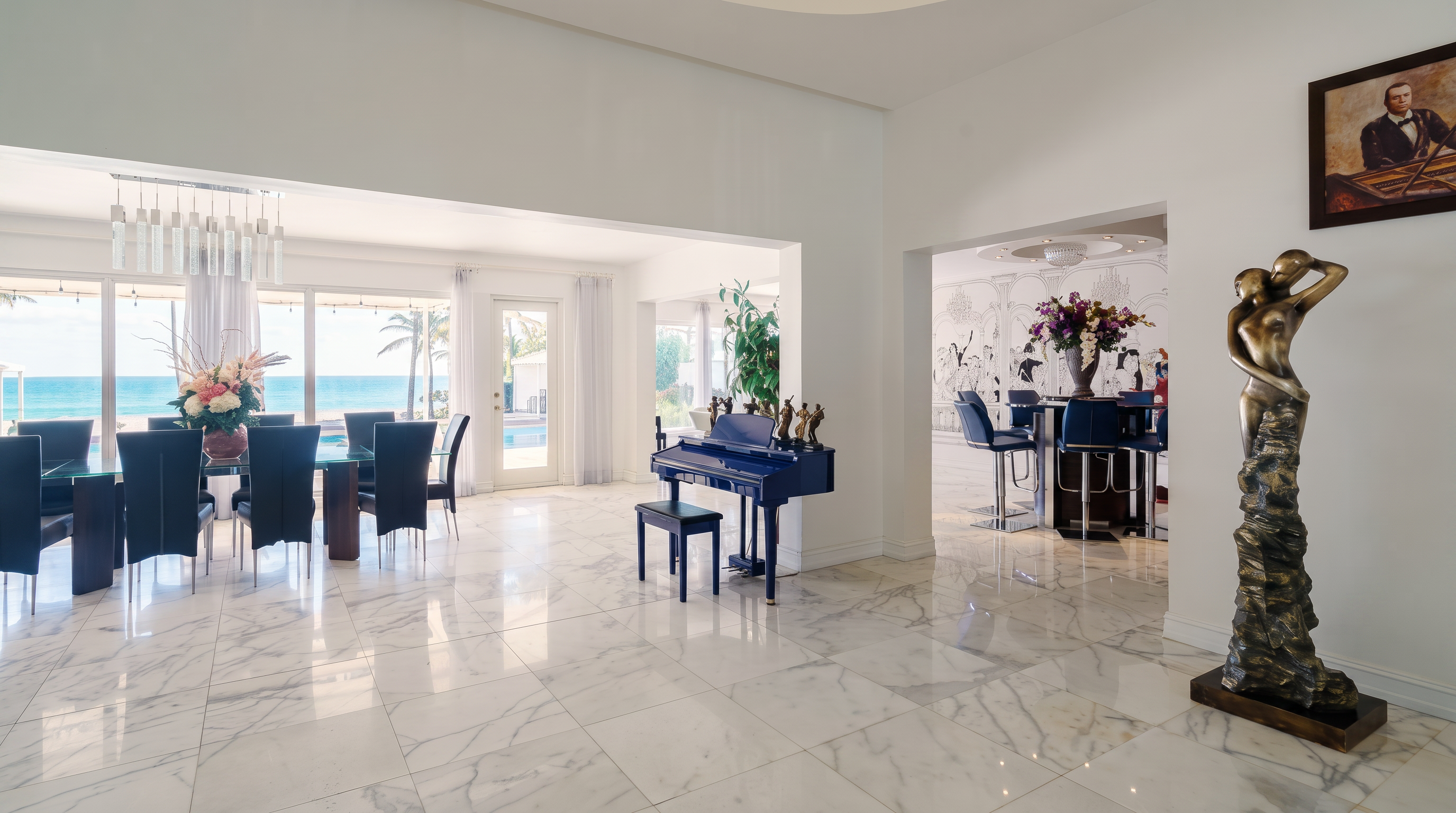 This spacious, open-concept interior features a formal dining area with a large glass-topped table and dark chairs, positioned near expansive windows that offer a clear view of the ocean. A striking blue piano sits in the center of the room, serving as a focal point, while a tall, abstract bronze sculpture stands prominently in the foreground. The space is finished with polished light-colored stone flooring and transitions into a secondary seating area with a bar, creating an elegant and airy atmosphere.