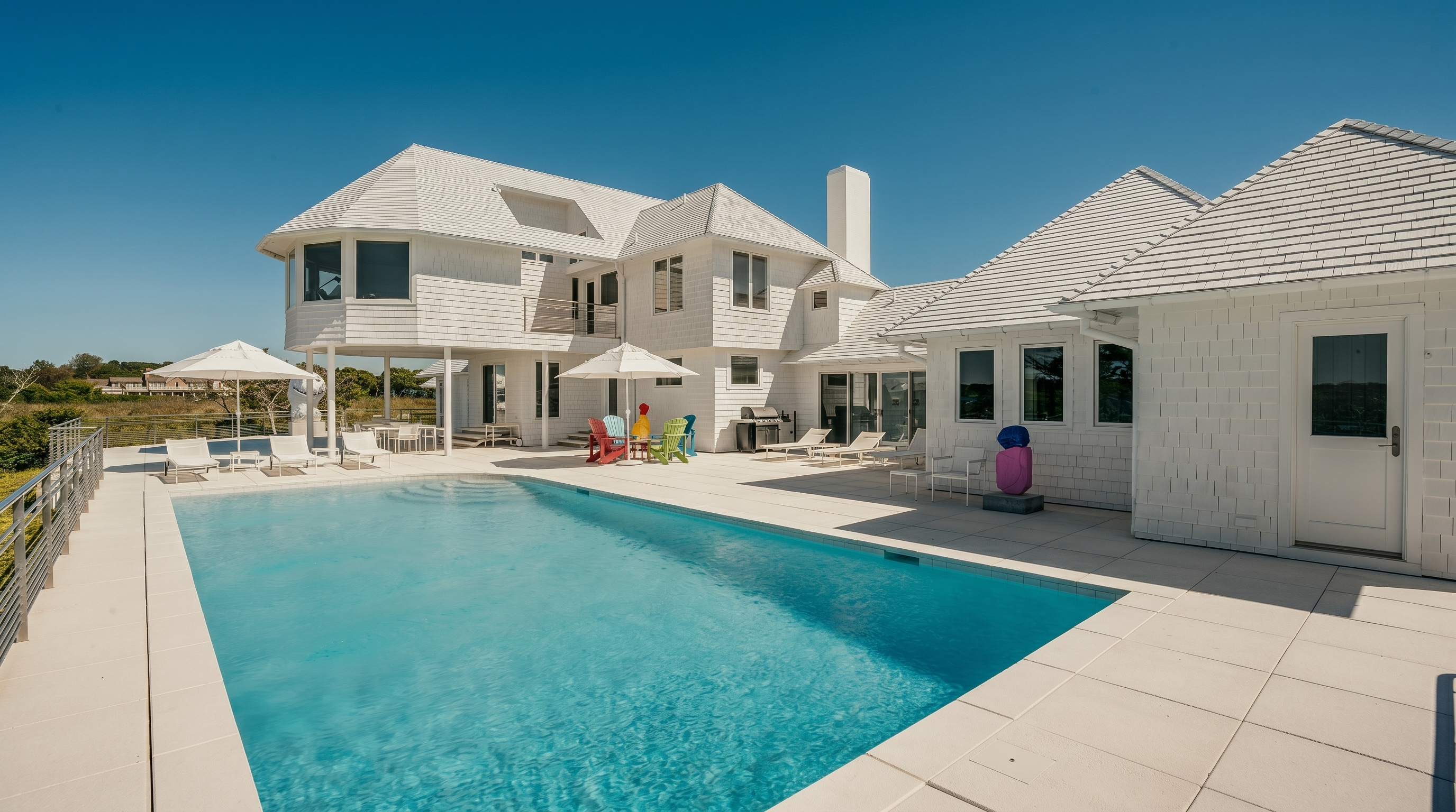 This bright, sun-drenched exterior view showcases a large, rectangular swimming pool set within a spacious, light-toned stone patio. The multi-level white shingled house features a classic coastal aesthetic with numerous windows and a prominent chimney, creating a clean and airy architectural profile. Several colorful Adirondack chairs and lounge seating areas are arranged around the pool, emphasizing a relaxed, resort-like atmosphere perfect for outdoor entertaining.