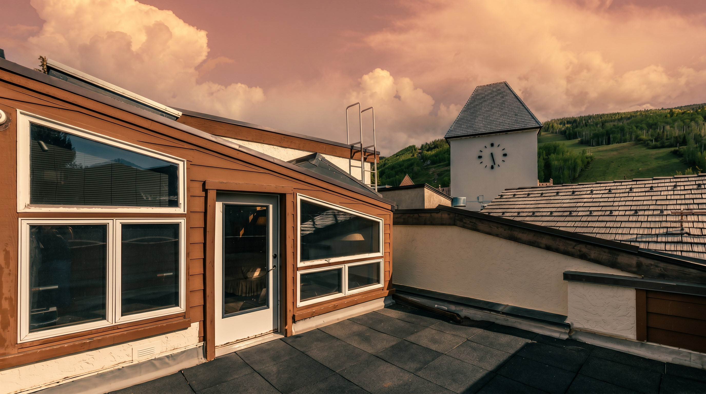 This image captures a rooftop deck featuring dark, square-tiled flooring and a brown-trimmed structure with large, multi-pane windows and a glass-paneled door. The perspective looks out toward a scenic mountain landscape, highlighted by a prominent clock tower building in the mid-ground. The scene conveys a sense of elevated, quiet outdoor living with a clear view of the surrounding alpine environment.