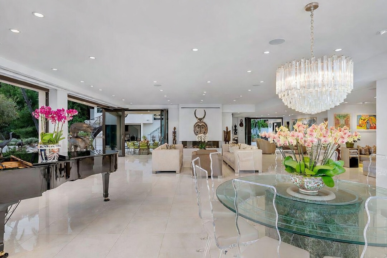 This expansive, open-concept living area features polished light-colored tile flooring and a grand crystal chandelier centered above a glass-topped dining table. The space is furnished with neutral-toned sofas arranged around a central fireplace with a tall, decorative surround, while a black grand piano sits prominently in the foreground. Large, retractable glass doors open the room to an outdoor patio, creating a seamless transition between the sophisticated interior and the exterior landscape.