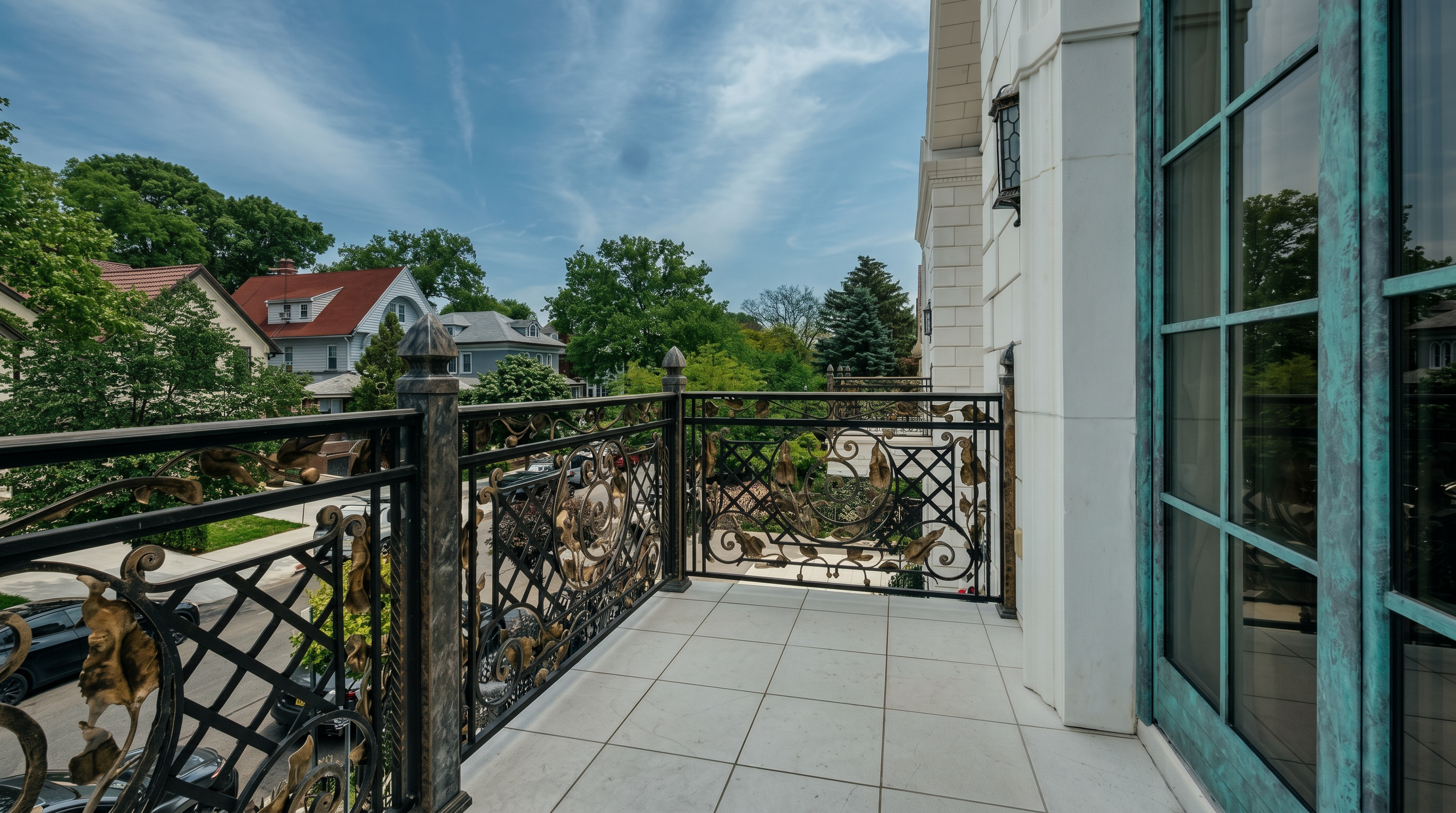 This elegant balcony features intricate, ornate wrought-iron railings with gold-leaf accents, providing a sophisticated and luxurious aesthetic. The space is finished with clean, light-colored square tiles and offers a clear view of the surrounding neighborhood and lush green trees. The perspective is from the balcony looking outward, highlighting the seamless transition between the interior living space and the outdoor area.