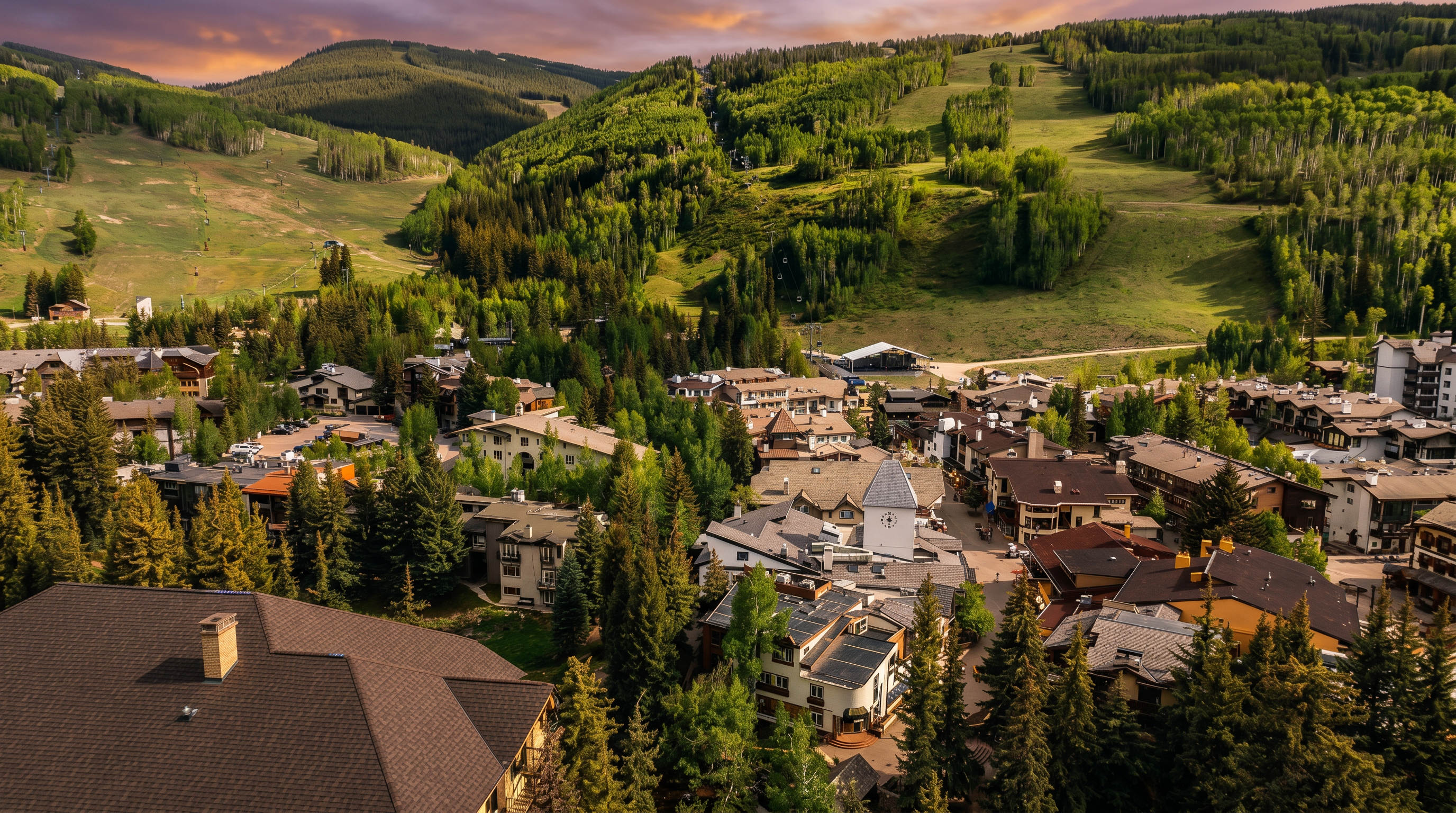 This high-angle aerial view captures a sprawling mountain resort village nestled at the base of a lush, green ski slope. The scene features a dense collection of multi-story residential buildings with varied rooflines, surrounded by mature pine and aspen trees. The perspective emphasizes the integration of the architecture into the natural landscape, showcasing the proximity of the village to the expansive mountain terrain.
