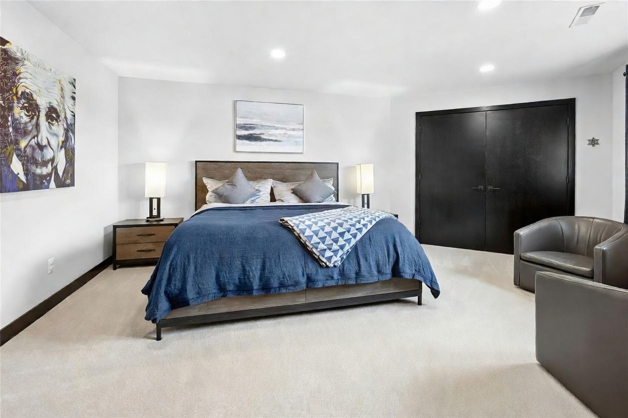 This spacious primary bedroom features a modern aesthetic with a central bed dressed in a deep blue comforter and a patterned throw. The room is accented by a large portrait of Albert Einstein on the left wall, a minimalist landscape painting above the headboard, and two sleek leather armchairs in the foreground. The neutral carpet and clean white walls create a bright, serene atmosphere, while the dark double doors add a sophisticated contrast.