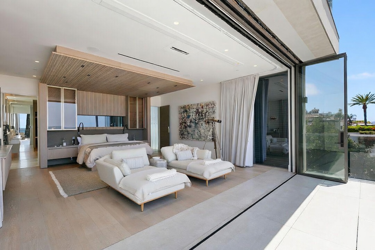 This expansive primary bedroom features a contemporary design with light wood paneling above the bed and a neutral color palette. The room is highlighted by large glass doors that slide open to reveal an outdoor terrace, seamlessly blending indoor and outdoor living spaces. Two white chaise lounges are positioned in the center of the room, facing the view, while a piece of abstract art adds a vibrant touch to the wall.