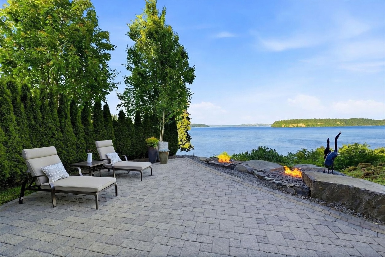 This serene outdoor patio features a paved stone surface furnished with two lounge chairs overlooking a expansive waterfront view. A low stone retaining wall incorporates built-in fire features, creating a cozy atmosphere against the backdrop of the bay and distant shoreline. The space is framed by a row of tall, manicured evergreen hedges and a slender tree, offering a private and tranquil setting for relaxation.