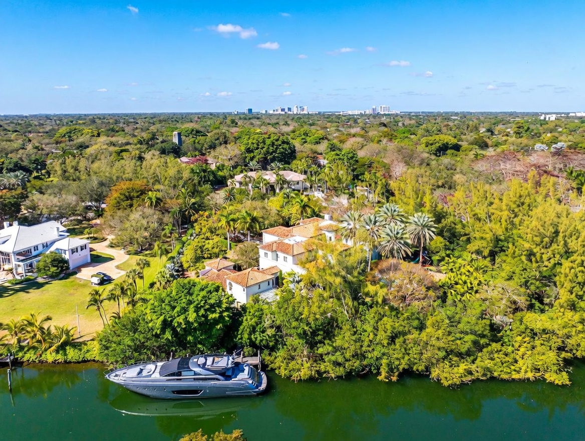 This high-angle aerial view captures a sprawling waterfront estate nestled within a lush, tropical landscape. The property features a multi-level residence with a classic tile roof, surrounded by dense greenery and palm trees, with a large private yacht docked along the canal edge. The perspective provides a comprehensive look at the expansive grounds and the serene, natural setting of the home.