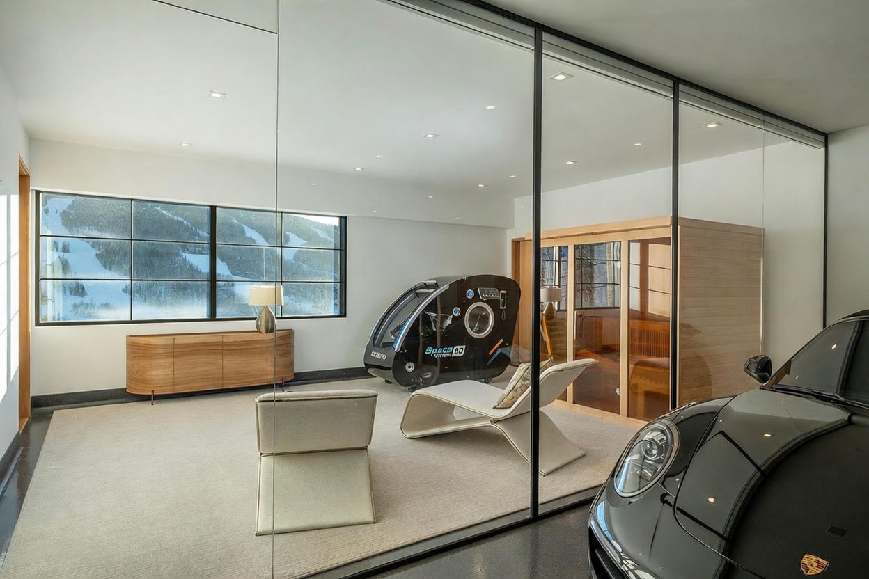 This modern, glass-enclosed home gym features a sleek, minimalist aesthetic with a view of a snowy mountain landscape through large windows. The space is equipped with a specialized oxygen therapy chamber, a contemporary lounge chair, and a wooden sauna, all set against a neutral carpeted floor. A portion of a black luxury vehicle is visible in the foreground, suggesting a unique integration between a garage and a wellness area.