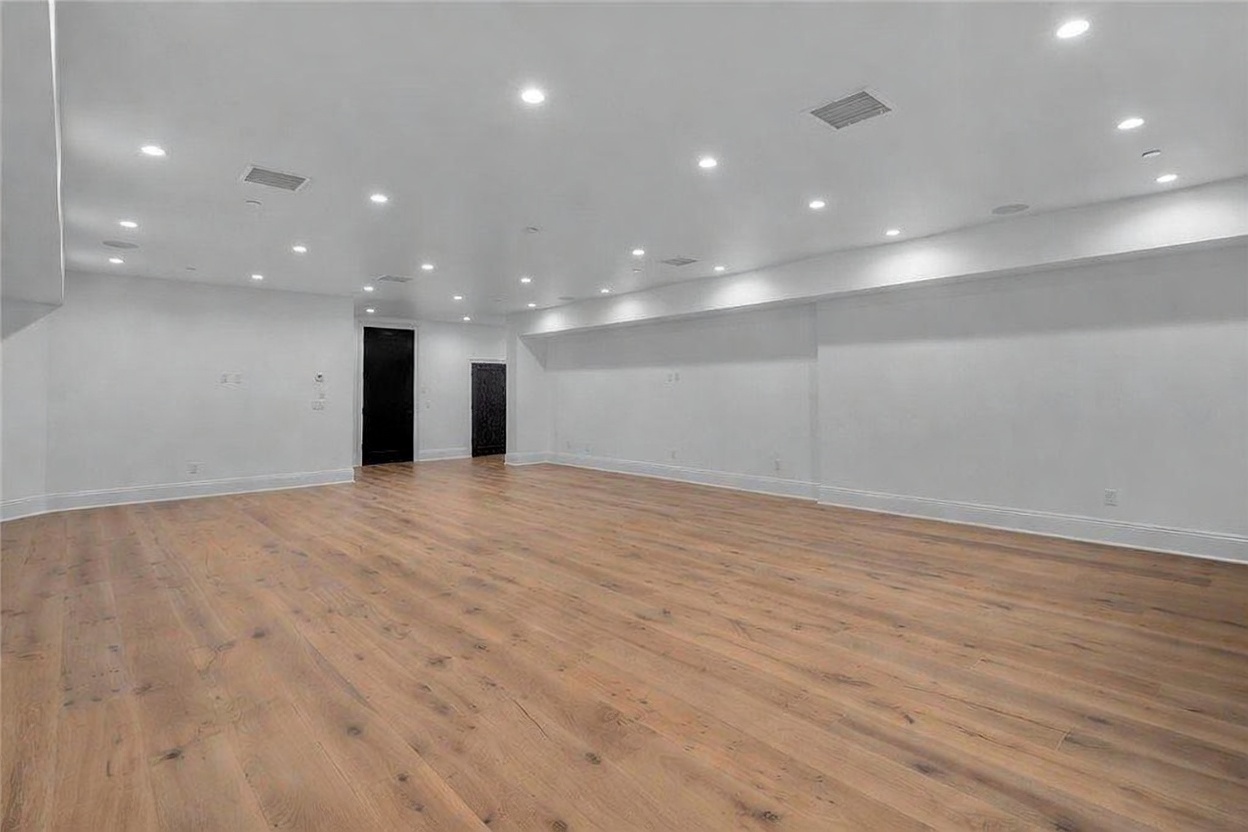 This spacious, open-concept basement features light-toned wood-look flooring and crisp white walls, creating a bright and airy atmosphere. The room is illuminated by numerous recessed ceiling lights and includes two dark-colored doors leading to adjacent areas. The perspective is wide and unobstructed, highlighting the expansive floor plan and clean, modern aesthetic.