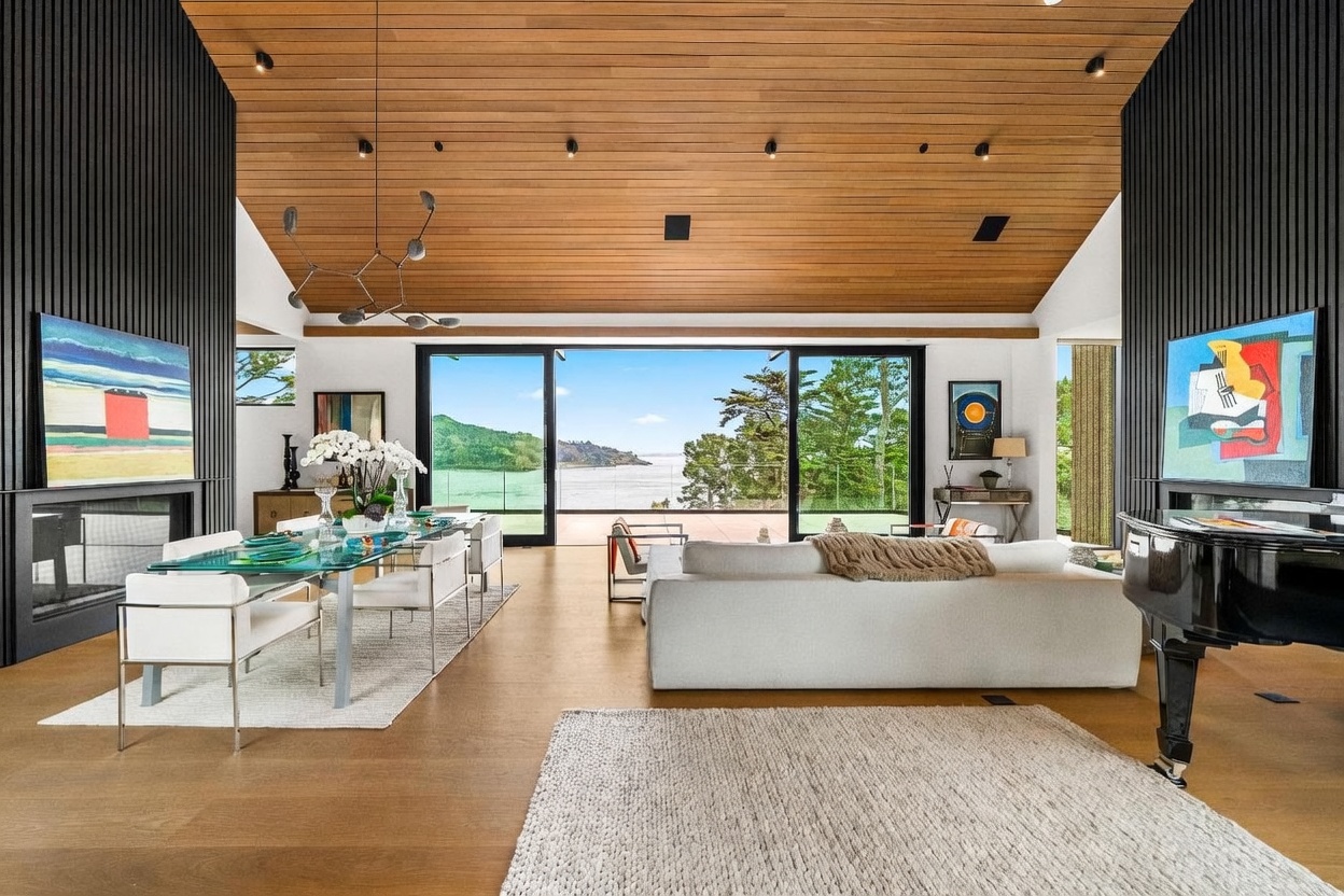 This expansive, open-concept living and dining area features a vaulted wood-paneled ceiling and light-toned wood flooring. A large white sofa faces a wall of glass sliders that open to a deck with panoramic water views, while a grand piano sits to the right. The space is defined by modern art, a sleek glass-topped dining table, and dark vertical wood paneling on the side walls, creating a sophisticated and airy atmosphere.