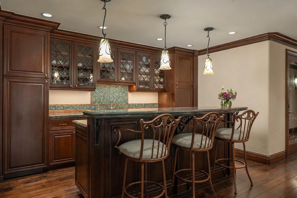 This elegant home bar area features rich, dark wood cabinetry with glass-front upper cabinets and a decorative green and cream tiled backsplash. A stone-topped bar counter provides seating for three with ornate metal bar stools, illuminated by three hanging pendant lights. The space is finished with dark wood flooring and crown molding, creating a sophisticated and inviting atmosphere.
