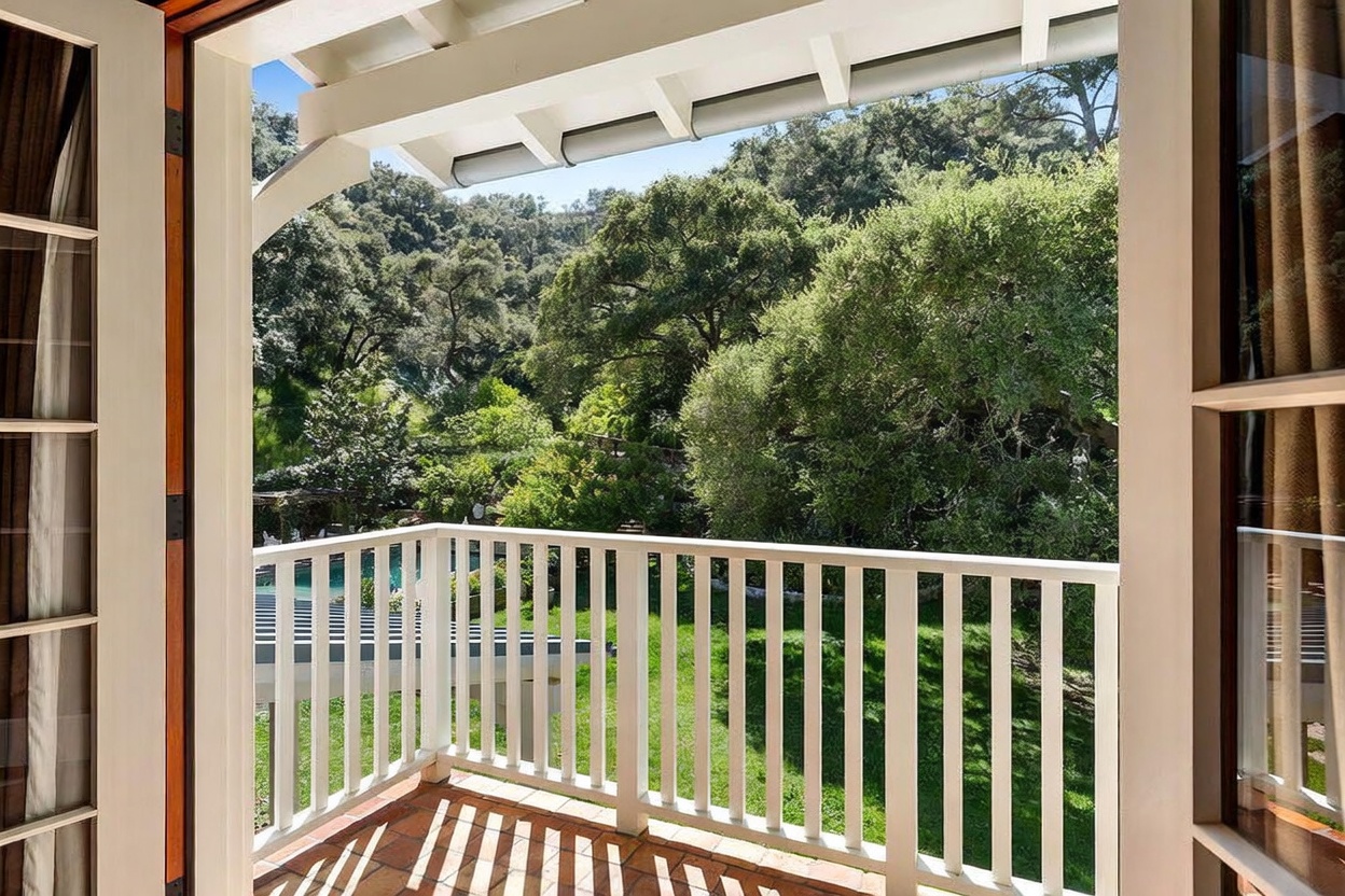 This image captures a view from a covered balcony looking out over a lush, tree-filled landscape. The balcony features a white railing and terracotta-toned flooring, framed by the interior door frame of the home. The perspective provides a serene, elevated vantage point that emphasizes the property's connection to the surrounding natural environment.