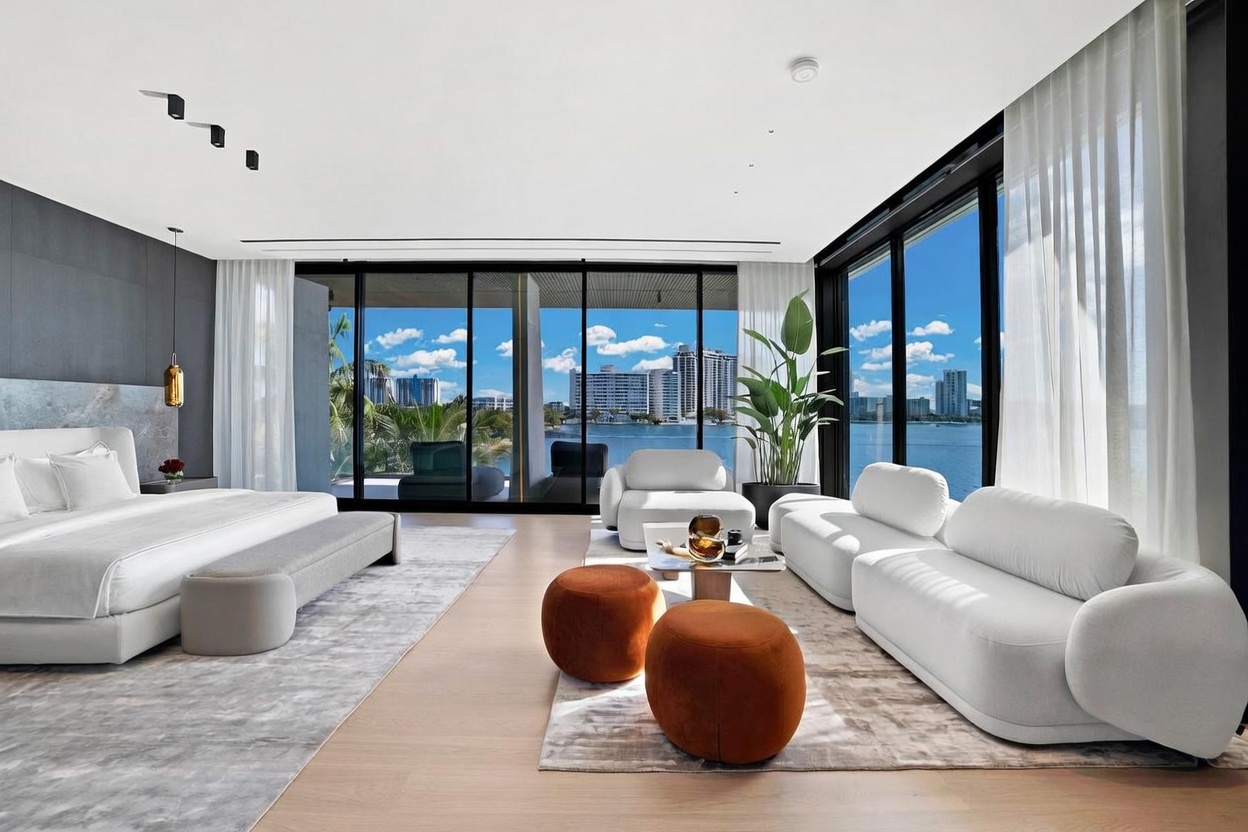 This expansive primary bedroom features a contemporary design with light wood flooring and a neutral color palette. The room is anchored by a large bed with a grey upholstered bench and is complemented by a comfortable seating area with white sofas and two round orange ottomans. Floor-to-ceiling windows provide an unobstructed, panoramic view of the waterfront and city skyline, creating a bright and airy atmosphere.
