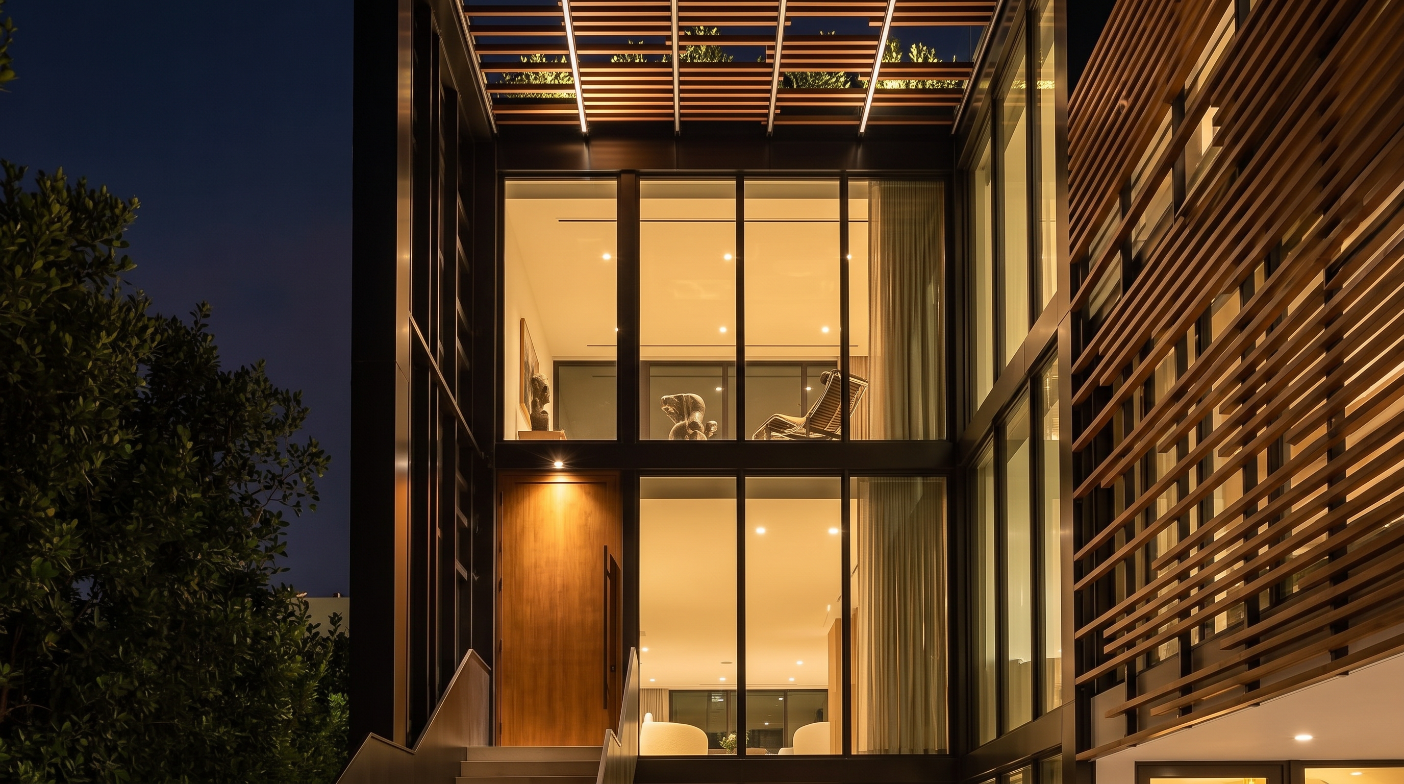 This modern, architectural entryway is characterized by a striking combination of dark steel framing, warm wood slats, and floor-to-ceiling glass. The house features a prominent solid wood front door leading up a sleek set of steps, illuminated by ambient evening lighting that highlights an elegant sculpture inside. The design seamlessly blends industrial elements with natural textures to create a sophisticated and inviting facade.