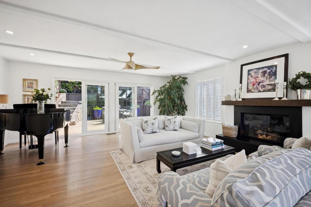 This bright and airy living room features light wood flooring, a neutral color palette, and a grand piano positioned near sliding glass doors that open to an outdoor deck. A comfortable white sofa faces a dark coffee table and a fireplace with a black surround and a wooden mantel, creating a cozy focal point. The space is accented by a large potted plant and a patterned area rug, offering a sophisticated and inviting atmosphere.