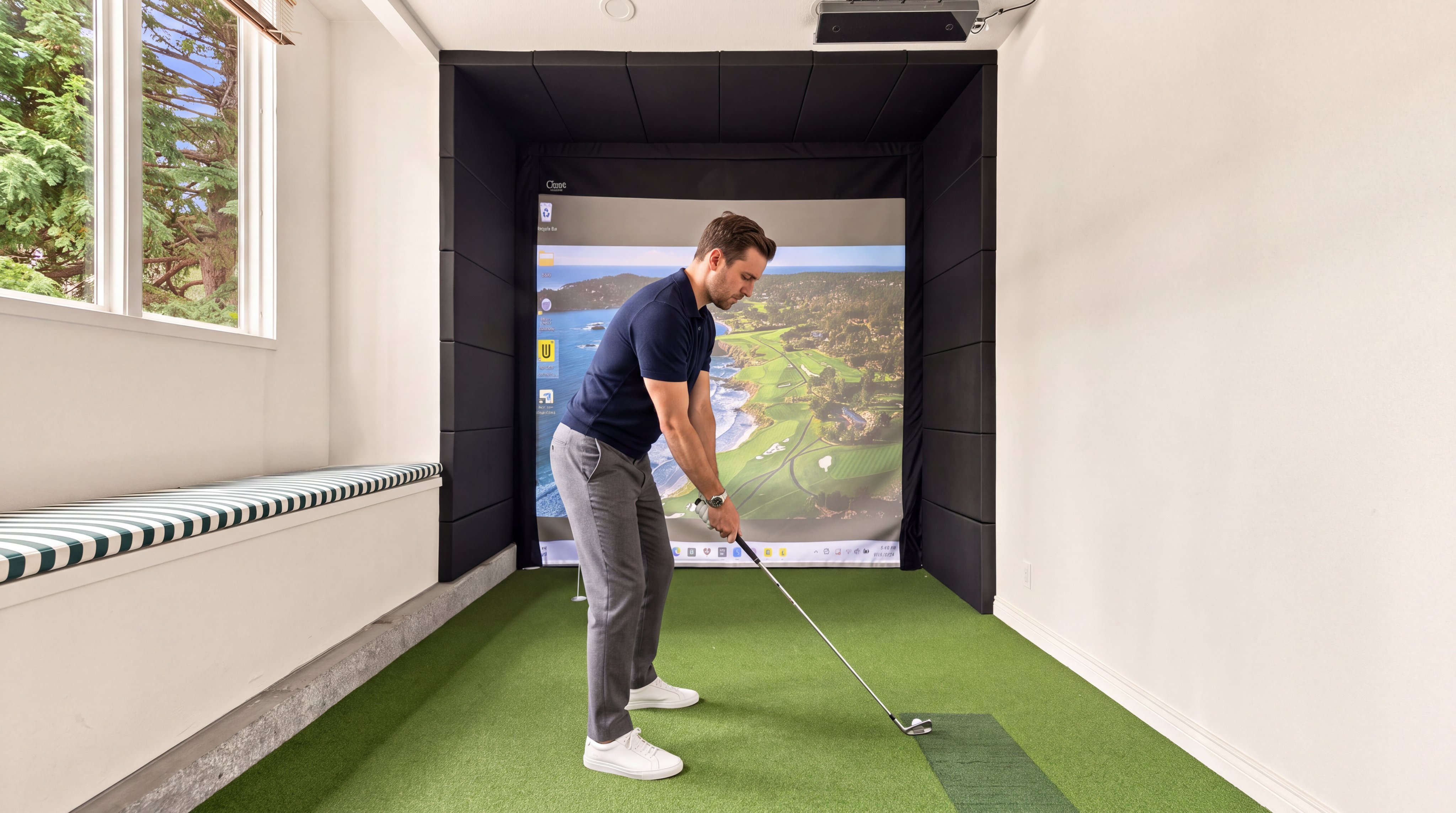 This interior space features a professional golf simulator setup with a green turf floor and a large projection screen displaying a scenic course. The room includes a built-in bench with patterned cushioning, clean white walls, and dark acoustic wall panels surrounding the simulator area. It provides a dedicated recreational space that is ideal for golf enthusiasts looking for indoor training.