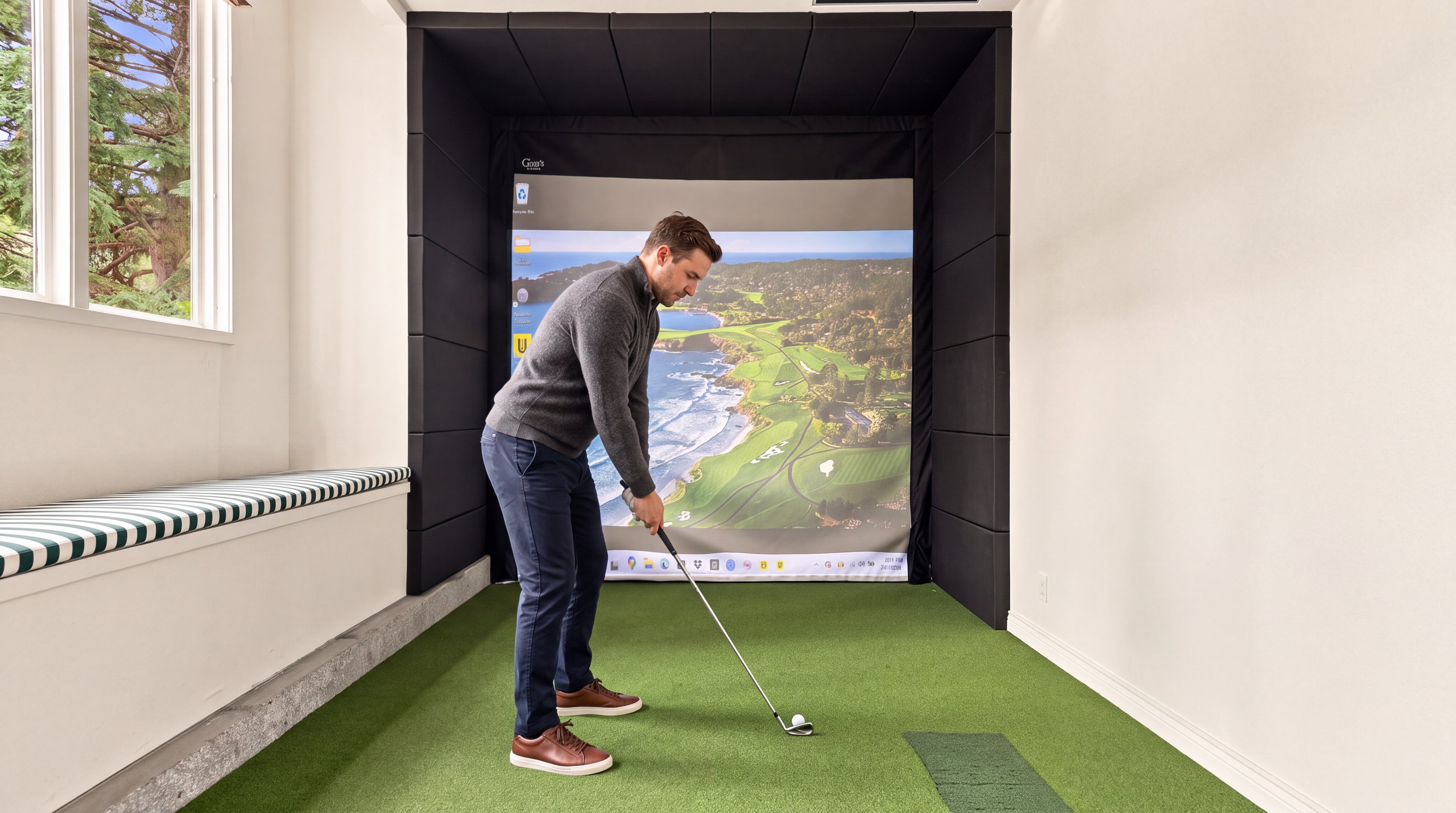 This interior space features a dedicated indoor golf simulator setup with a high-quality projection screen and artificial turf flooring. The room includes built-in black acoustic paneling surrounding the screen and a built-in bench seating area, offering a sophisticated and functional space for recreation and practice. The design combines utility with modern aesthetics, making it an attractive amenity for a luxury residence.