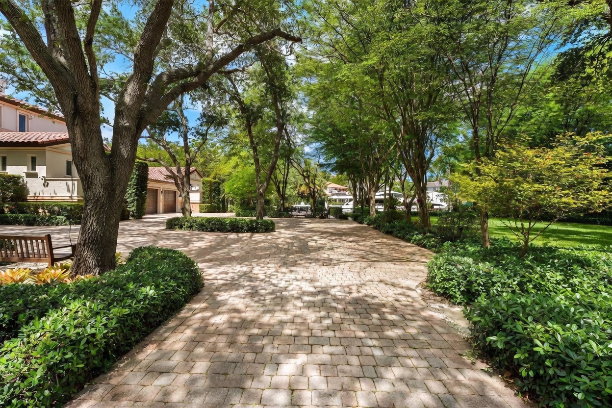 This image captures a serene, tree-lined driveway paved with light-colored stone pavers, leading toward a residential property. Mature trees provide a lush canopy over the path, creating a dappled light effect on the ground, while a wooden bench sits to the left under a large tree. The perspective is a ground-level view looking down the driveway, emphasizing the private, park-like atmosphere of the estate.