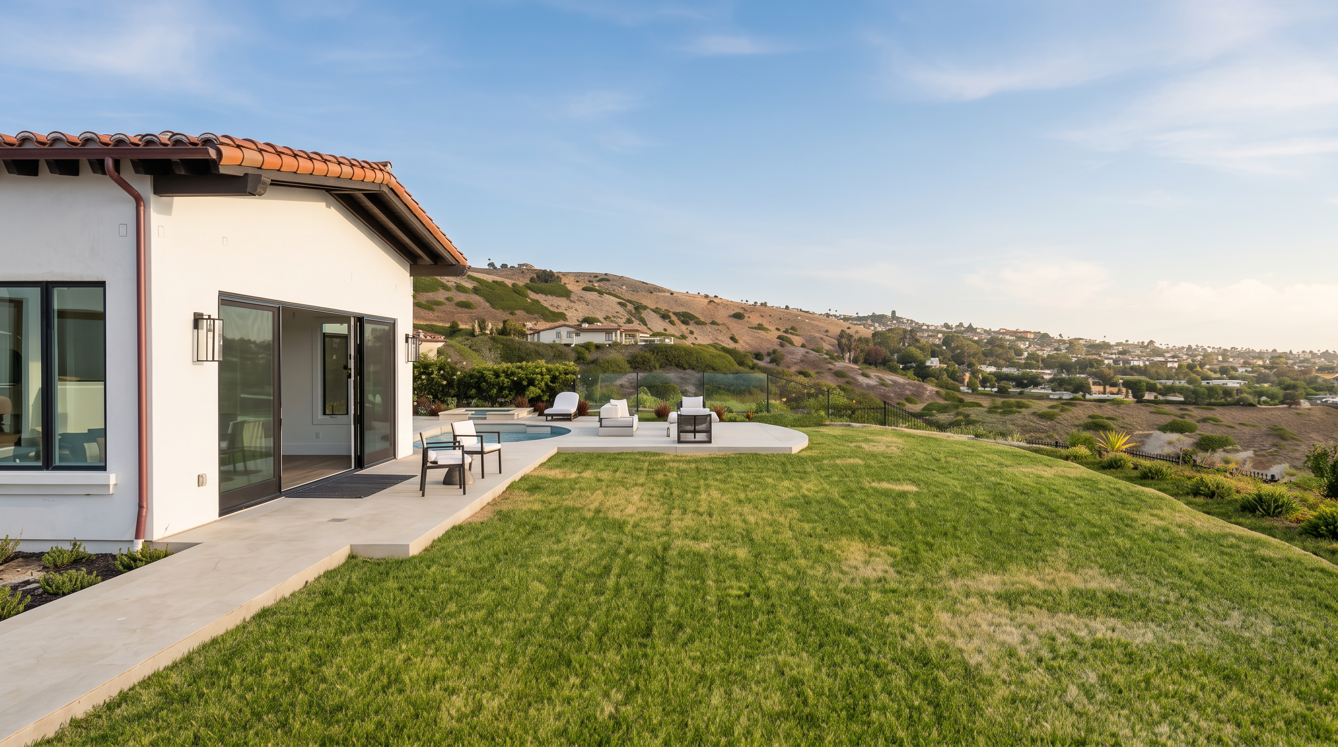 This rear view of a modern home showcases a seamless transition between indoor and outdoor living spaces, featuring large sliding glass doors that open onto a paved patio. The patio area includes a swimming pool and lounge chairs, overlooking a vast, rolling hillside landscape. The bright, sunny atmosphere highlights the expansive lawn and the home's clean, Mediterranean-inspired architectural style.