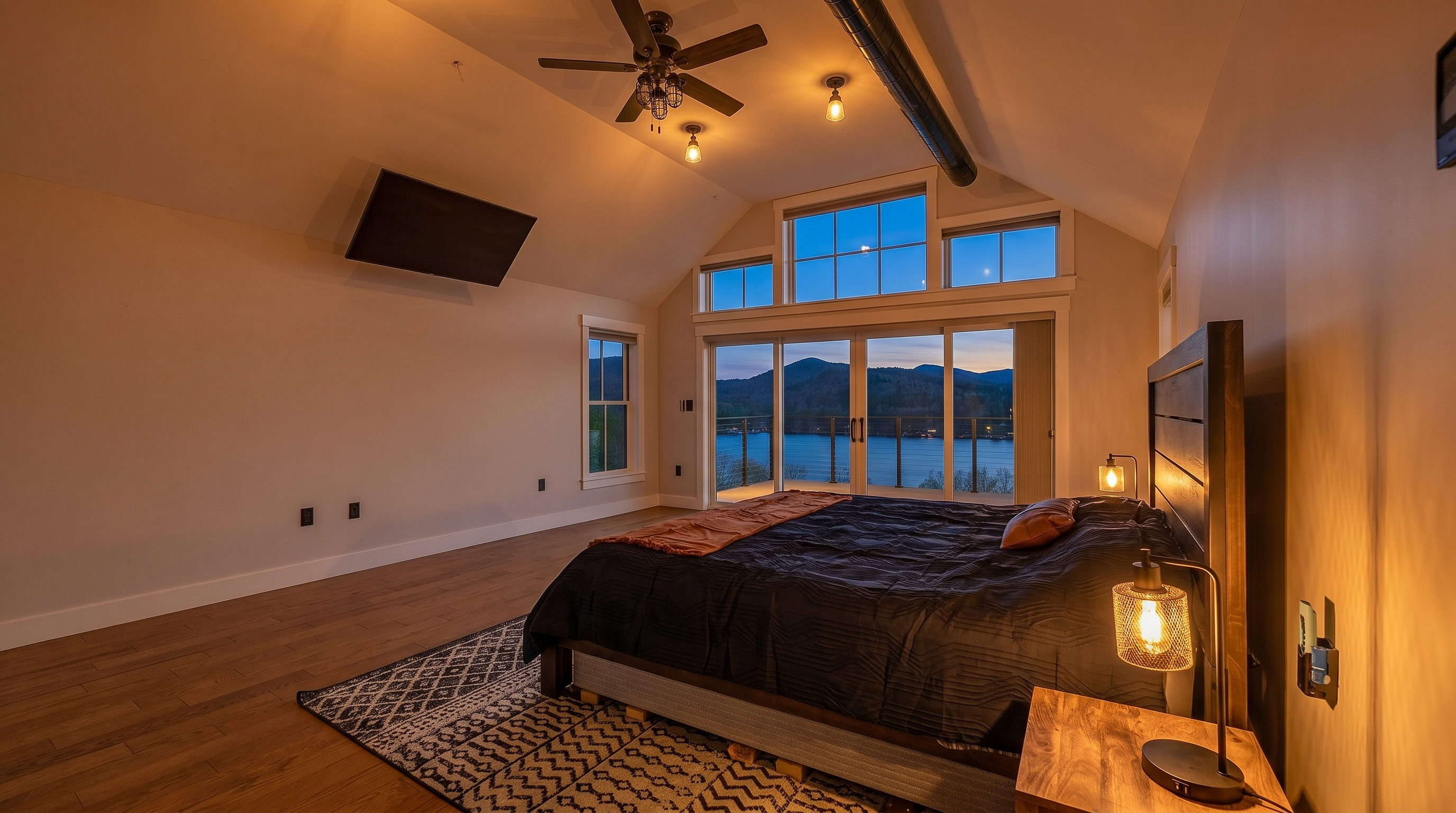 This spacious primary bedroom features vaulted ceilings with exposed ductwork and a ceiling fan, creating a modern, industrial-inspired aesthetic. A large bed sits atop a patterned area rug, positioned to take advantage of the expansive windows and sliding glass doors that offer panoramic views of the lake and mountains. The room is finished with light-toned walls and warm wood-look flooring, providing a bright and airy atmosphere.