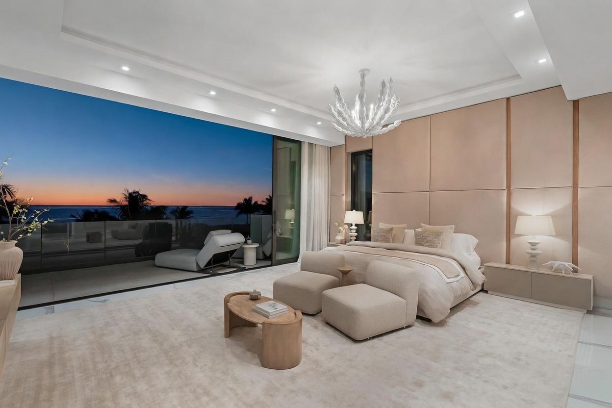 This luxurious primary bedroom features a neutral, monochromatic palette with soft textures and a large bed as the focal point. A striking, sculptural chandelier hangs above the room, while floor-to-ceiling glass doors open onto a private balcony with expansive ocean views at sunset. The space is elegantly appointed with two modern armchairs, a low-profile coffee table, and custom wall paneling that adds depth and sophistication to the serene environment.