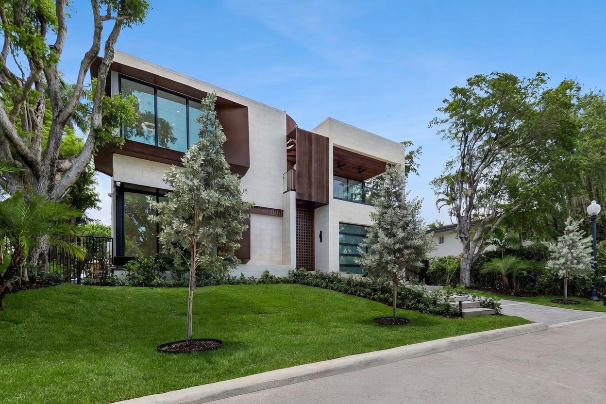 This modern, multi-level residence features a striking architectural design with a mix of light-colored stone cladding and dark, metallic-toned accents. The front facade is defined by large, expansive windows and a recessed entryway, set against a manicured lawn with mature trees. The perspective is a low-angle street view, emphasizing the home's contemporary aesthetic and clean, geometric lines.