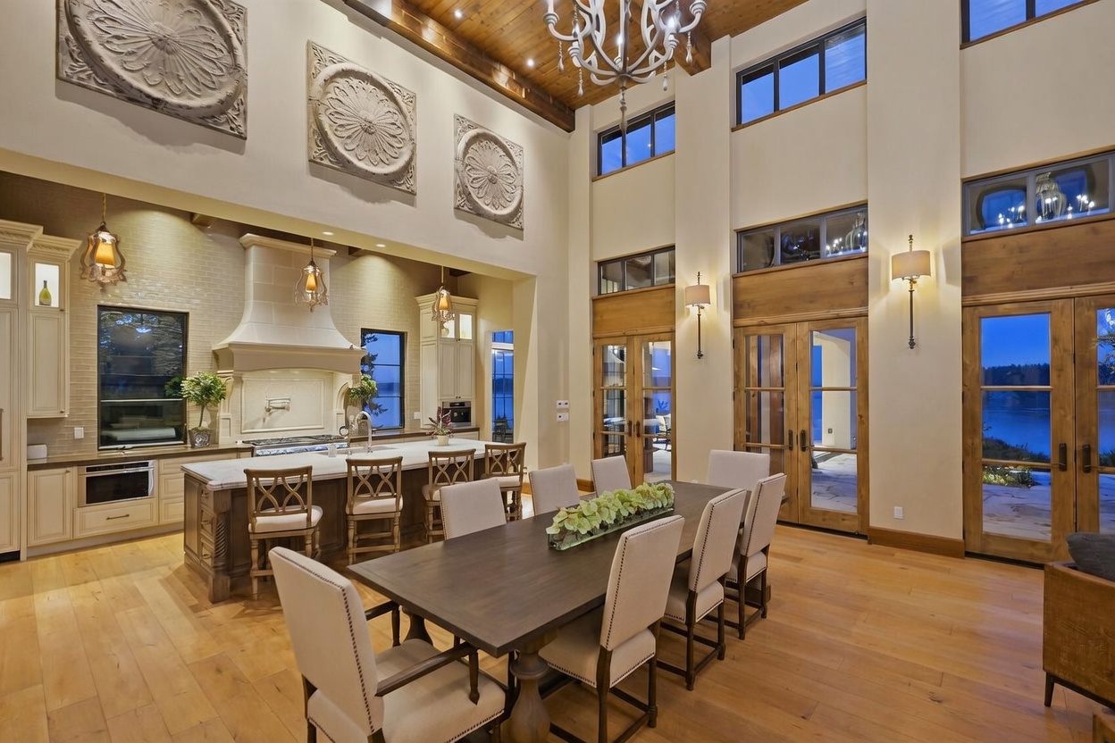 This expansive open-concept space features a large kitchen with a central island and a formal dining area, all under a high, wood-beamed ceiling. The room is adorned with decorative wall medallions and illuminated by a grand chandelier, while tall windows and glass doors provide views of the outdoors. The aesthetic combines rustic wood elements with elegant, neutral-toned furnishings to create a sophisticated and airy atmosphere.