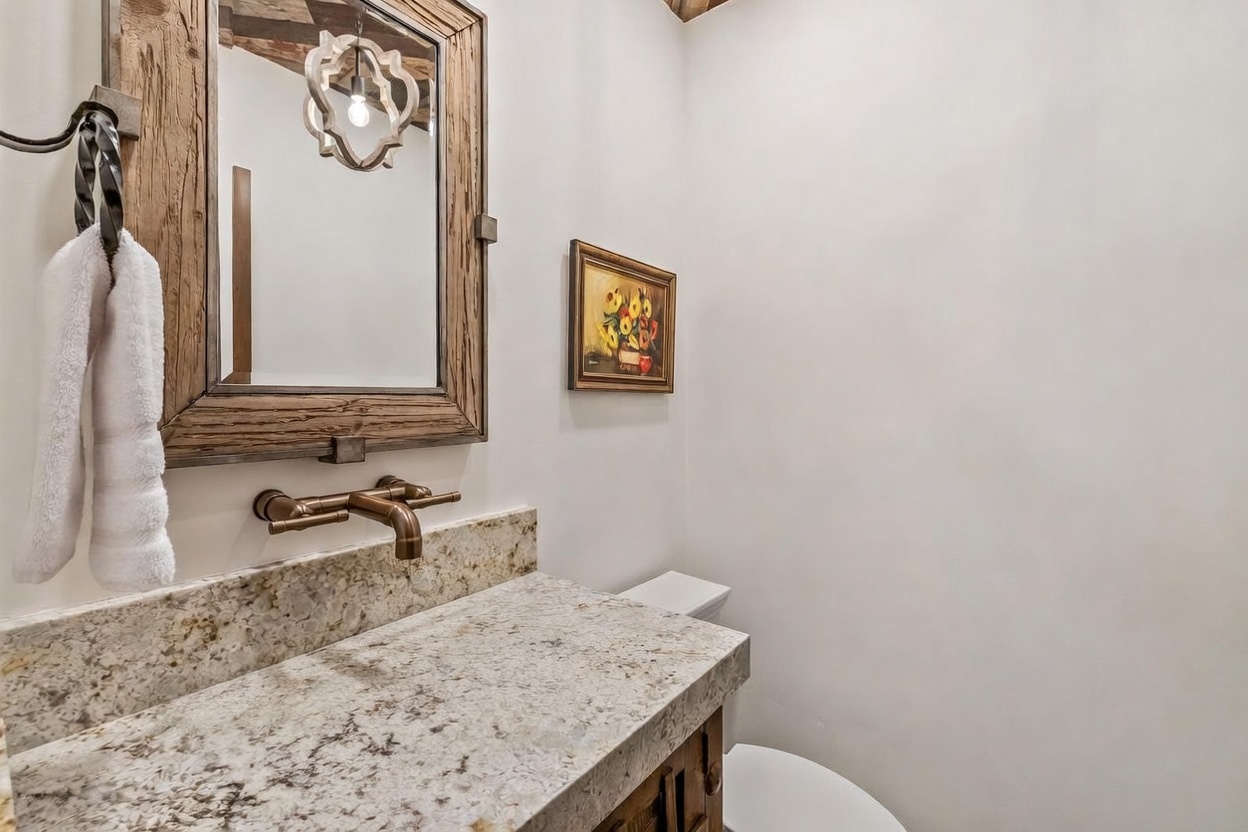This rustic-style powder room features a distinctive hammered metal vessel sink set into a light-colored stone countertop. A wooden-framed mirror hangs above the vanity, complemented by a wall-mounted bronze faucet and a framed floral painting on the adjacent wall. The space is finished with clean, neutral walls and a white toilet, creating a warm and inviting atmosphere.
