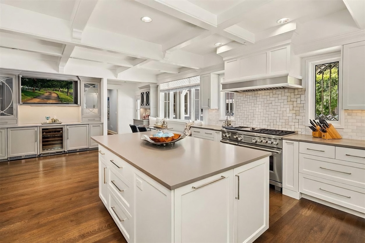 This bright and spacious kitchen features a large central island with a solid-surface countertop and white cabinetry. The space is equipped with a professional-style range, a tiled backsplash, and a built-in wine refrigerator integrated into the cabinetry. The room is accented by coffered ceilings and warm wood flooring, creating an inviting and high-end atmosphere.