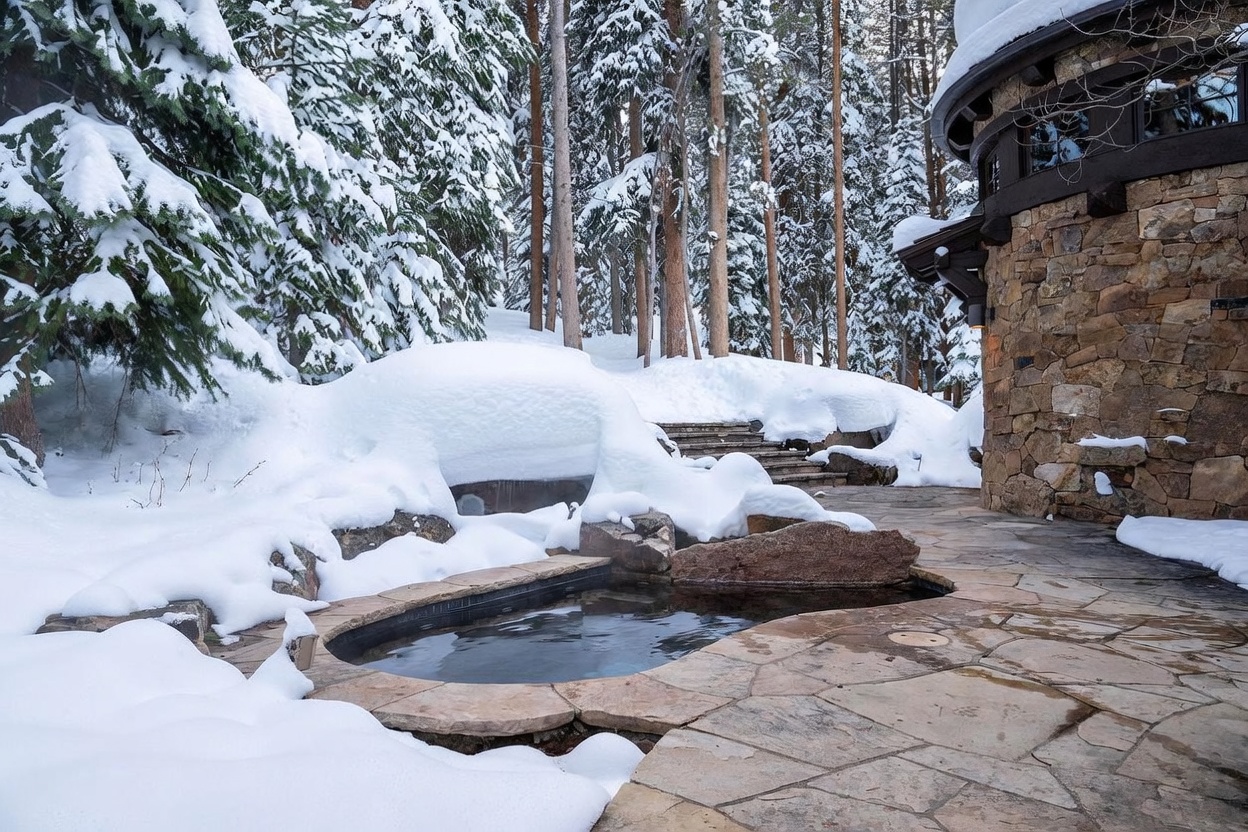 This image captures a serene outdoor spa area nestled in a snowy, wooded landscape. A stone-lined hot tub sits flush with a flagstone patio, adjacent to a rustic stone-clad building with dark-framed windows. The scene is framed by snow-covered pine trees and a gentle stone staircase leading up the hillside, creating a tranquil and luxurious winter retreat atmosphere.