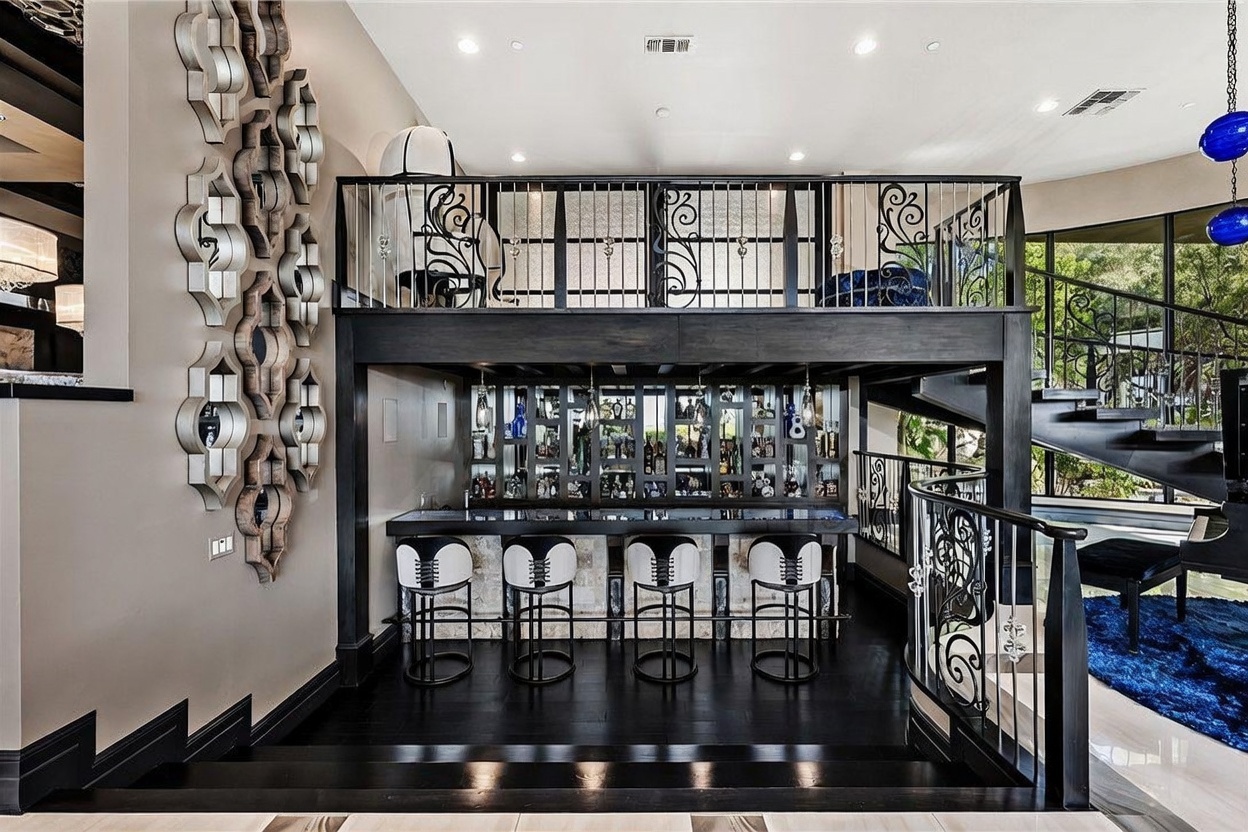 This sophisticated interior space features a custom-built, two-story home bar area with a dark wood finish and integrated shelving for glassware and spirits. A mezzanine level with decorative metal railings overlooks the bar, which is complemented by four modern bar stools and a polished dark floor. To the left, a large, ornate wall sculpture adds a dramatic artistic element, while a staircase with a matching metal railing leads to the upper level.