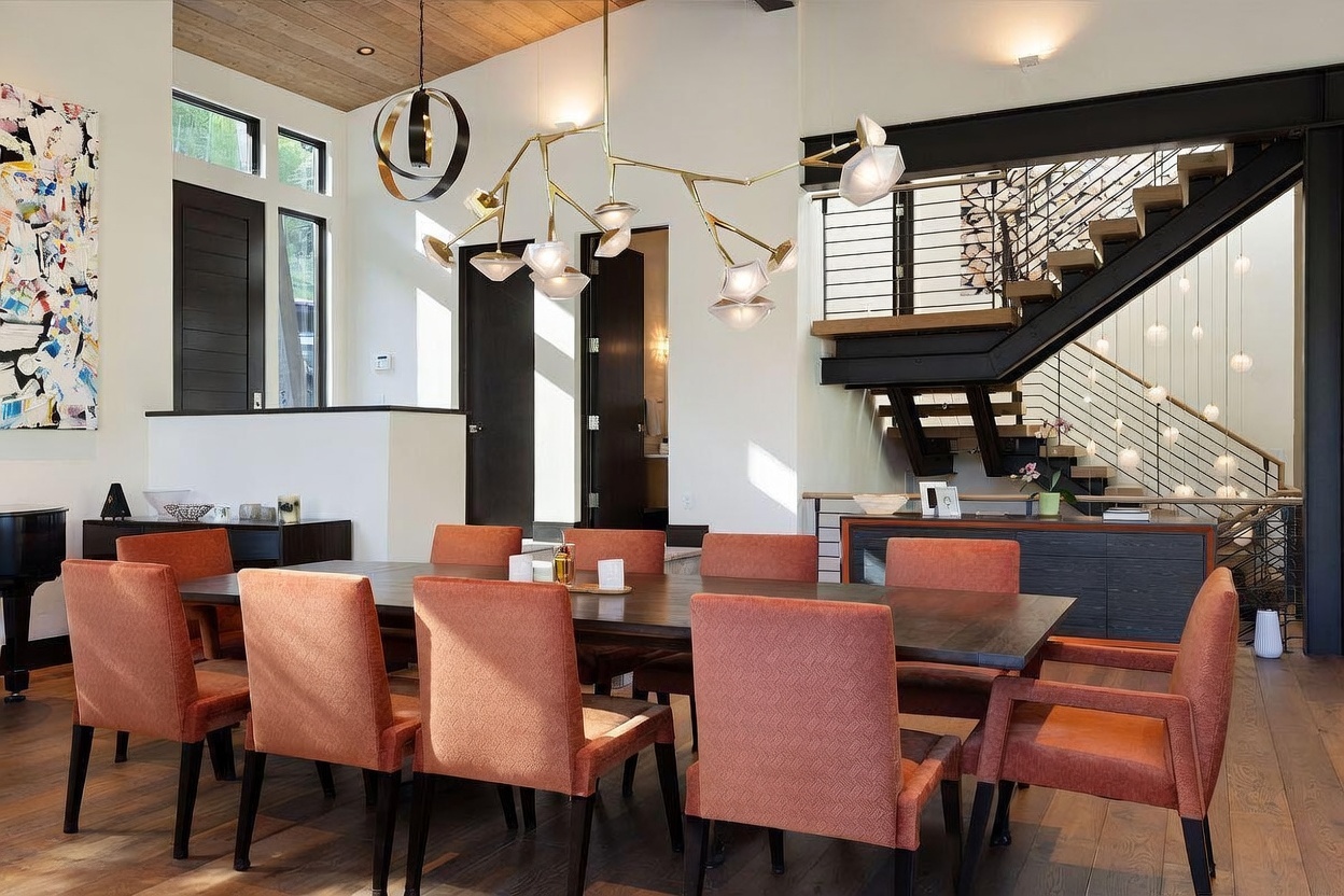 This elegant dining room features a large, dark wood table surrounded by eight upholstered chairs in a warm, burnt-orange hue. A striking, multi-armed modern chandelier serves as the focal point above the table, while a staircase with a metal railing and a cascading light fixture creates a sophisticated backdrop. The space is characterized by high ceilings, clean lines, and a blend of dark wood and neutral wall tones, offering a refined and inviting atmosphere.