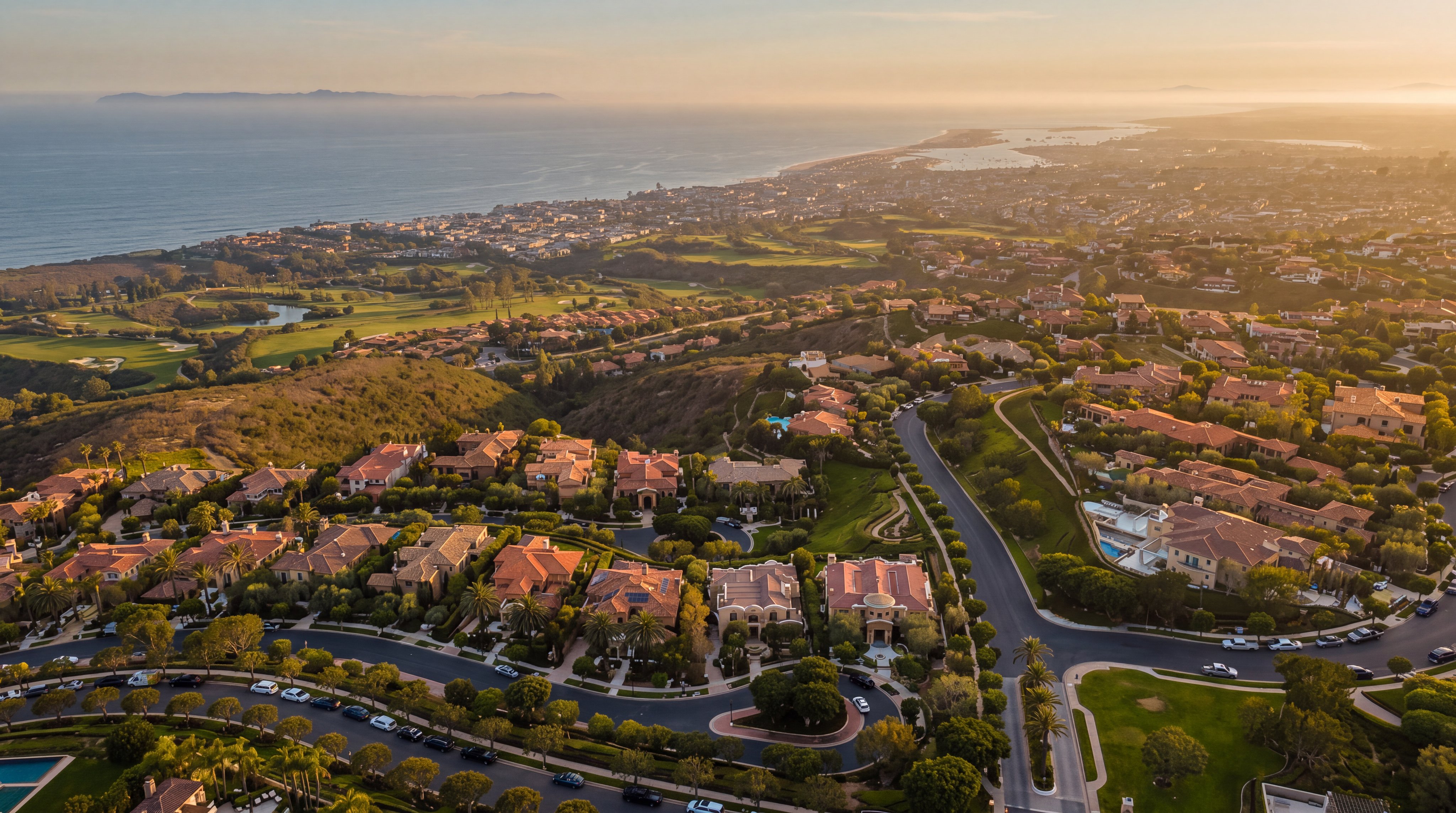 This high-altitude aerial view captures a sprawling, upscale residential community nestled along a coastal hillside. The perspective highlights the property at 3 Island Vista, showcasing its prominent position within the neighborhood, surrounded by lush landscaping, winding roads, and a nearby golf course. In the background, the expansive blue ocean stretches toward the horizon, emphasizing the prime location and scenic coastal setting.