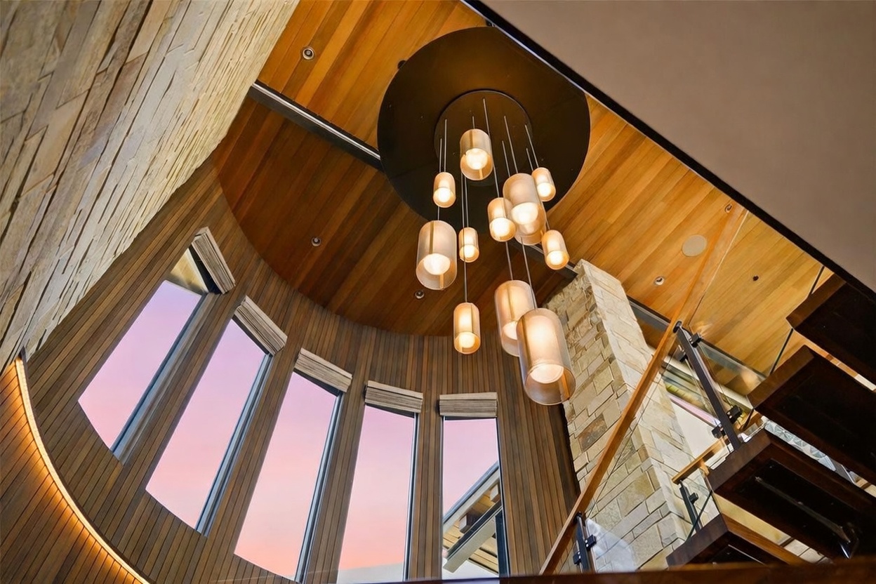 This dramatic interior view captures a multi-story stairwell featuring a striking, tiered chandelier with cylindrical glass pendants suspended from a circular mount. The space is defined by warm wood-paneled ceilings and curved walls, complemented by a tall, textured stone-clad chimney breast. Large, curved windows allow natural light to filter in, highlighting the rich wood tones and the modern architectural design of the staircase.