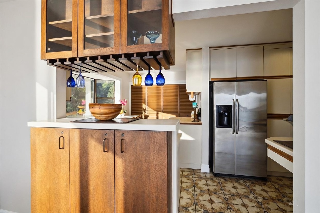 This kitchen features a warm, mid-century aesthetic with wood cabinetry and a hanging glass rack above a peninsula counter. The space includes a stainless steel refrigerator, patterned tile flooring, and a window that allows natural light to filter into the room. The perspective captures the transition between the breakfast bar area and the main kitchen workspace.