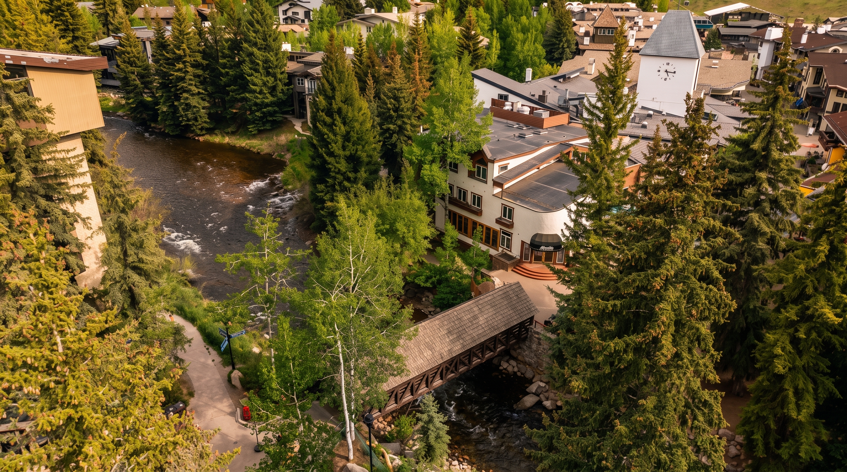 This high-angle aerial view captures a charming mountain resort setting, featuring a wooden covered bridge spanning a flowing creek. The scene is surrounded by lush evergreen trees and multi-story buildings with traditional alpine-inspired architecture, including a prominent clock tower. The perspective highlights the integration of the natural landscape with the pedestrian-friendly village layout.