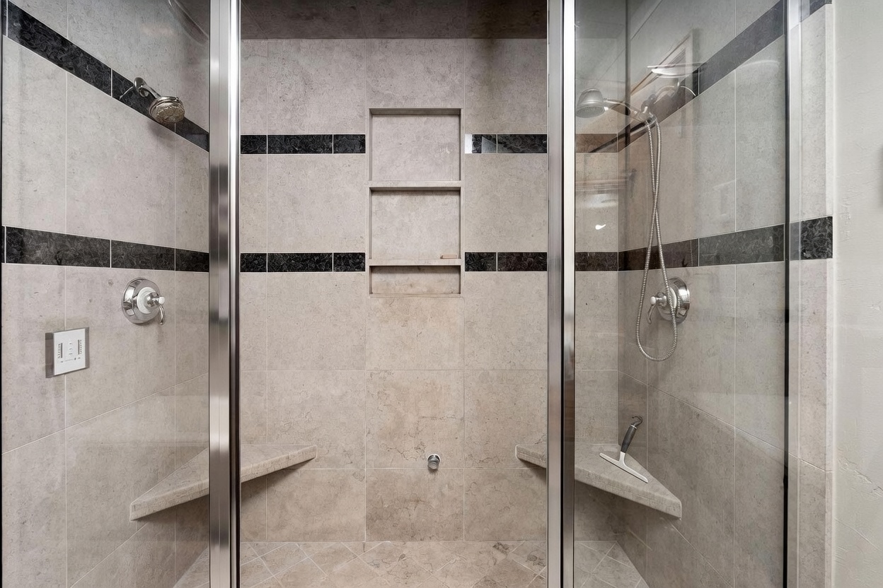 This spacious walk-in shower features a dual-head design with polished chrome fixtures and a central three-tiered recessed shelving unit. The walls are finished in large, neutral-toned stone tiles accented by horizontal bands of dark, polished stone, while matching corner benches provide convenient seating on both sides. The perspective is centered, looking directly into the glass-enclosed shower stall, highlighting its clean, symmetrical, and functional layout.