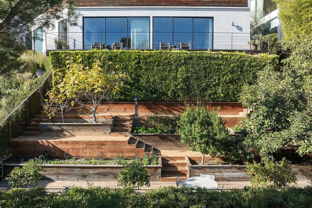 This multi-tiered terraced garden features raised wooden planter beds filled with lush greenery and small trees, creating a structured and functional outdoor space. A set of wooden stairs connects the levels, leading up to a modern home with a large balcony featuring glass railings and expansive windows. The scene is captured from an elevated perspective, highlighting the integration of tiered landscaping with the contemporary architecture of the residence.