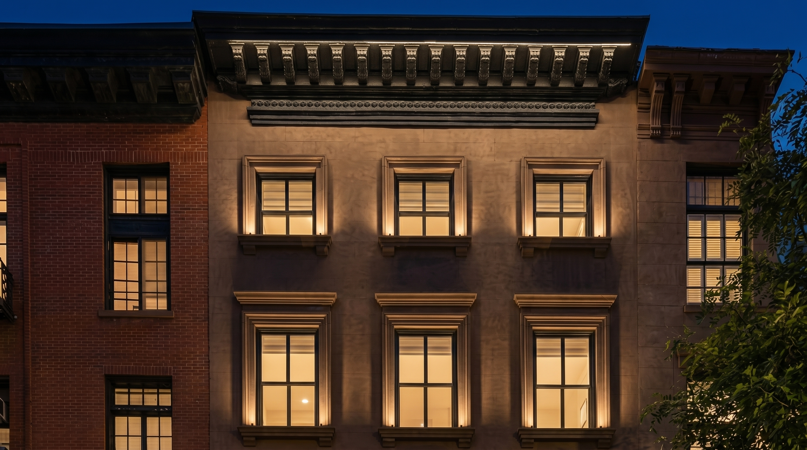 This image showcases the front facade of a classic NYC-style brownstone building, characterized by its symmetrical window placement and ornate architectural detailing. The building features sturdy, cornice-topped window frames and a robust, decorative roofline that reflects traditional urban architecture. The exterior brick and masonry work presents a well-maintained, historic aesthetic suitable for high-end residential living.