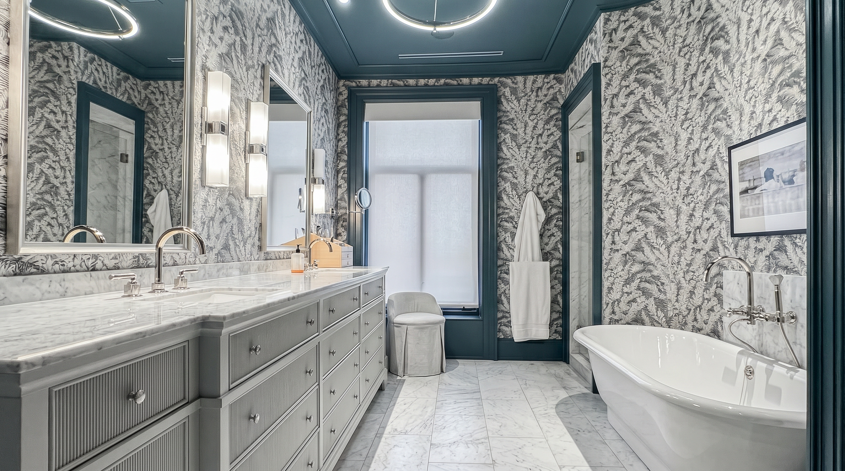 This luxurious primary bathroom features a long double vanity with stone countertops, patterned wallpaper, and a deep soaking tub positioned near a window. The room is defined by a striking deep blue ceiling and matching trim, which contrasts elegantly with the light-toned stone flooring. The perspective captures the full length of the space, highlighting the sophisticated lighting fixtures and the refined, high-end aesthetic of the design.
