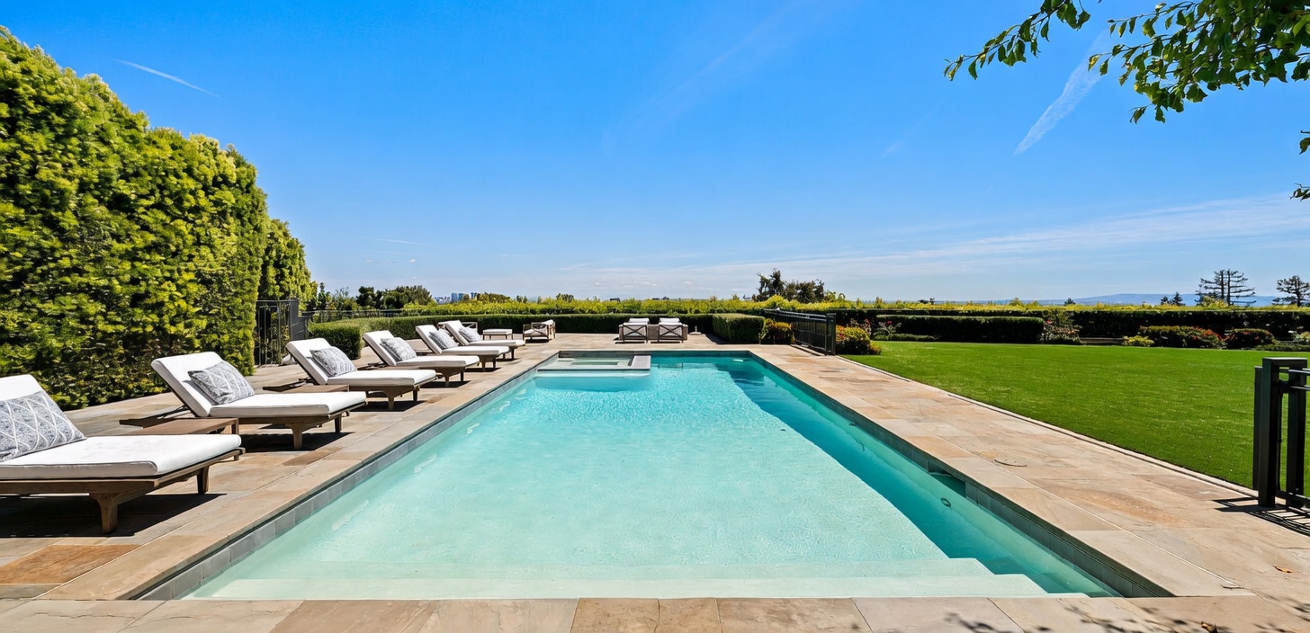 This expansive outdoor space features a rectangular swimming pool with an integrated spa, surrounded by a stone patio lined with multiple lounge chairs. A manicured green lawn stretches out to the right, bordered by neatly trimmed hedges that frame a clear, panoramic view of the horizon. The scene is captured from a low, eye-level perspective, emphasizing the clean lines of the pool and the serene, sunny atmosphere of the backyard.