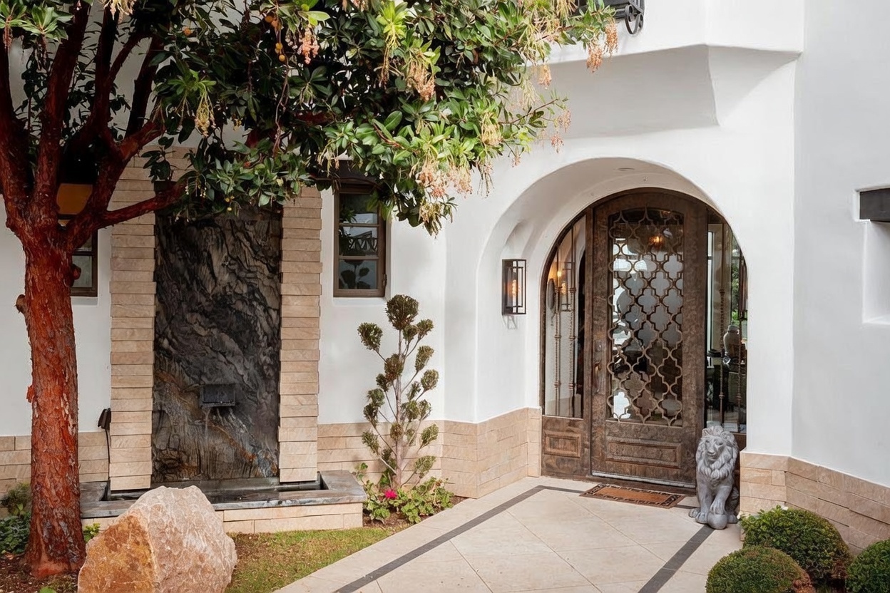 This inviting entryway features a grand arched doorway with an intricate metal grille, set against a crisp white stucco facade. To the left, a striking water feature with a dark, textured stone wall adds a sophisticated architectural element, complemented by a mature tree and manicured landscaping. The scene is completed by a stone-paved walkway and a decorative lion statue, creating a refined and welcoming curb appeal.