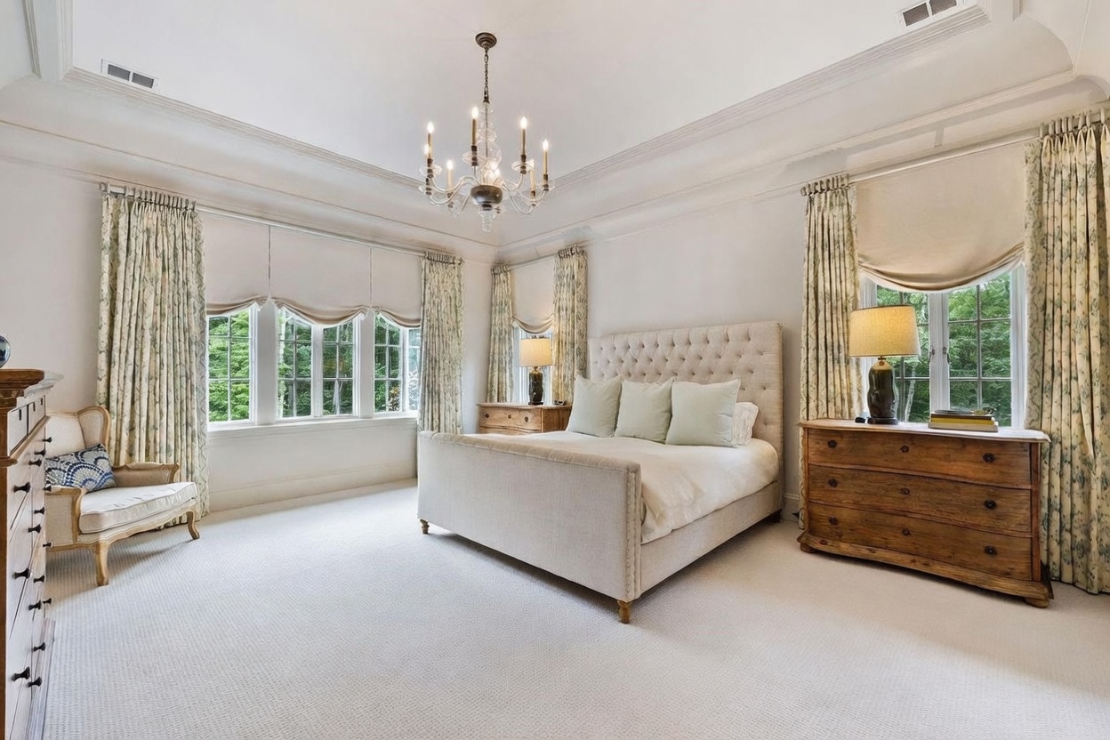 This elegant primary bedroom features a neutral color palette, a tufted upholstered bed, and a crystal chandelier centered in a tray ceiling. Large windows with patterned drapery and valances allow ample natural light to fill the space, while two wooden dressers serve as bedside tables. The room is finished with plush carpeting, creating a serene and sophisticated atmosphere.