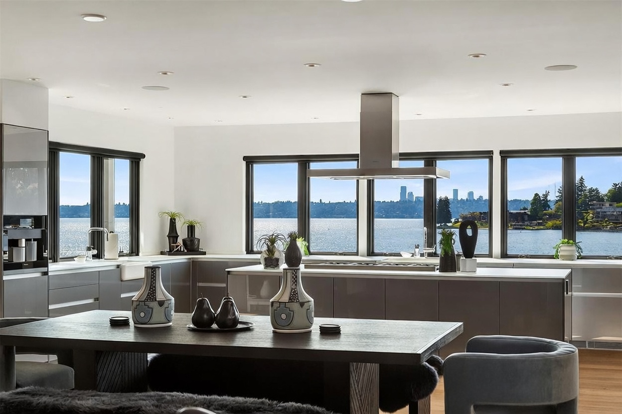 This modern, open-concept kitchen features sleek, dark-toned cabinetry and a large central island with a built-in cooktop and stainless steel range hood. A dining table sits in the foreground, while expansive windows provide a panoramic view of the water and distant city skyline. The space is bright and airy, characterized by a minimalist aesthetic and high-end finishes.
