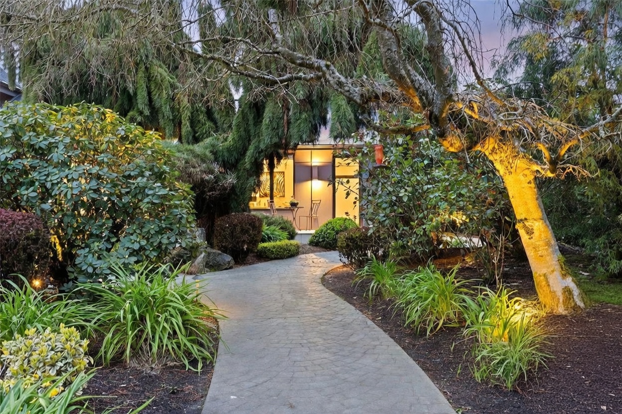 A beautifully landscaped garden path leads toward a modern, illuminated entryway, framed by lush greenery and mature trees. The scene is captured at dusk, with warm exterior lighting highlighting the textured stone walkway and the surrounding foliage. The overall impression is one of tranquility and inviting curb appeal, perfect for a serene residential setting.