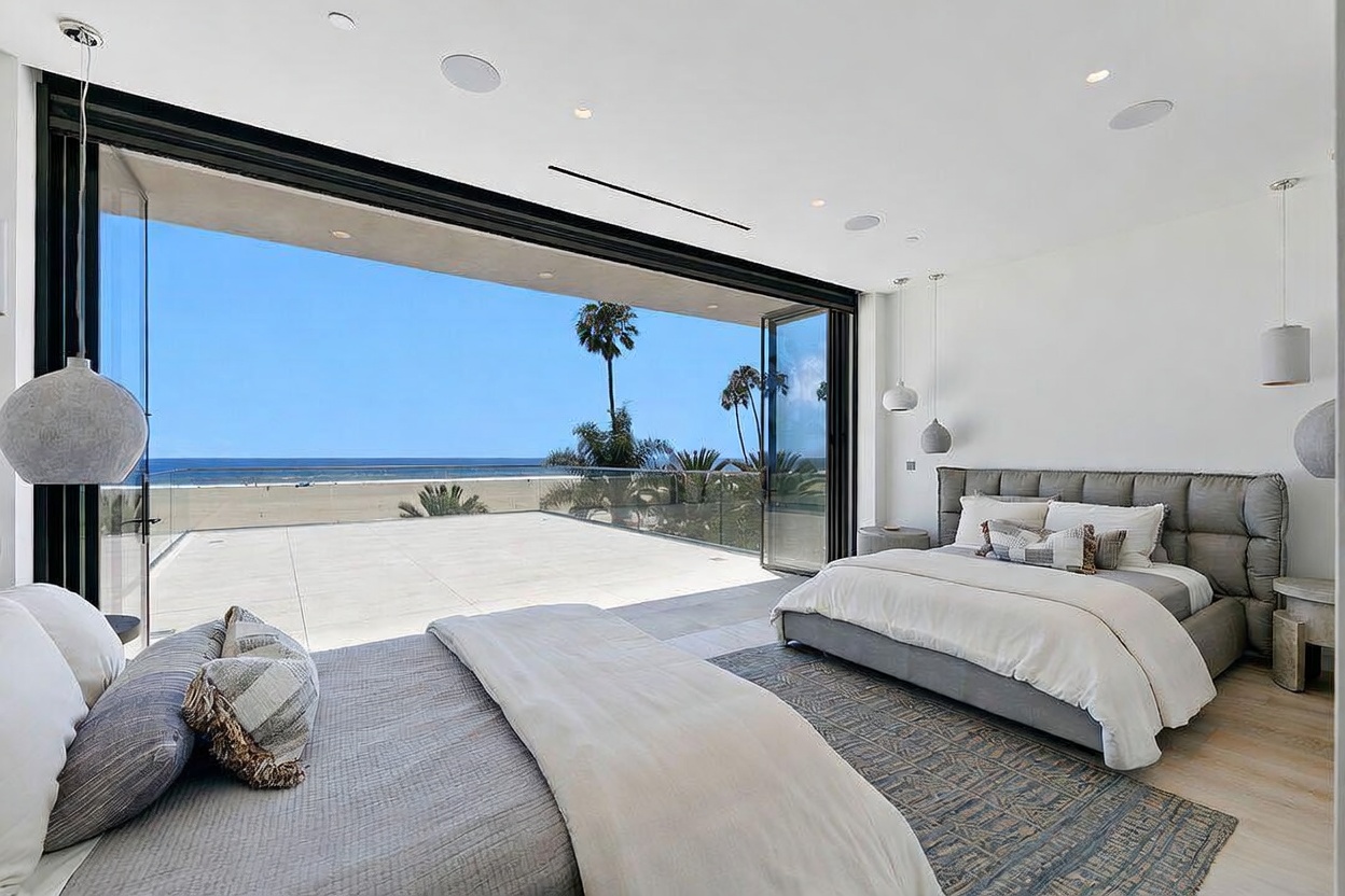 This expansive primary bedroom features a contemporary design with two beds and a neutral color palette. The room is defined by massive, floor-to-ceiling glass folding doors that open completely to a spacious balcony, providing an unobstructed, panoramic view of the ocean and beach. The minimalist aesthetic is complemented by soft pendant lighting and a textured area rug, creating a serene and luxurious atmosphere.