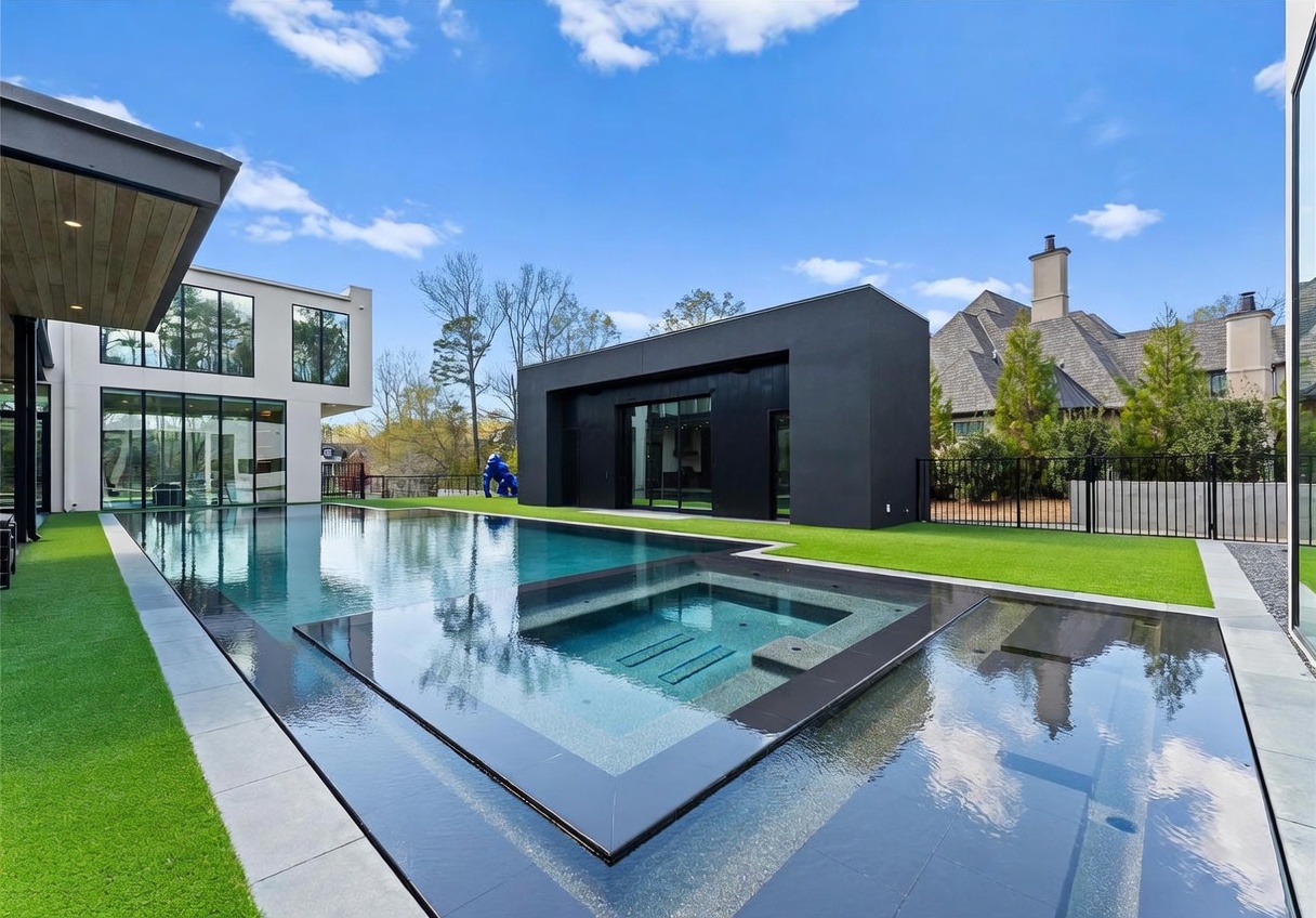 This striking modern backyard features a sleek, rectangular infinity-edge pool with an integrated spa, set against a backdrop of contemporary architecture. The property includes a minimalist black structure, expansive glass walls on the main residence, and a manicured artificial turf lawn. The perspective captures the clean lines and high-end design of the outdoor living space, emphasizing a luxurious and private atmosphere.