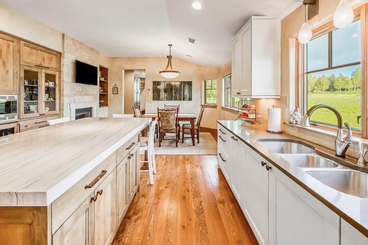 This bright, open-concept kitchen features a large central island with a light wood finish and a stone countertop, complemented by white cabinetry along the perimeter. A dining area is visible in the background, anchored by a wooden table and chairs, while a stone-clad chimney breast adds texture to the space. The room is bathed in natural light from large windows that offer a scenic view of the surrounding landscape.